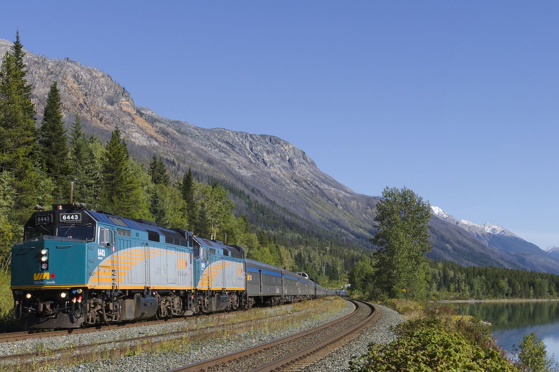 Train traveling along tracks near a lake and mountains, under a clear blue sky.