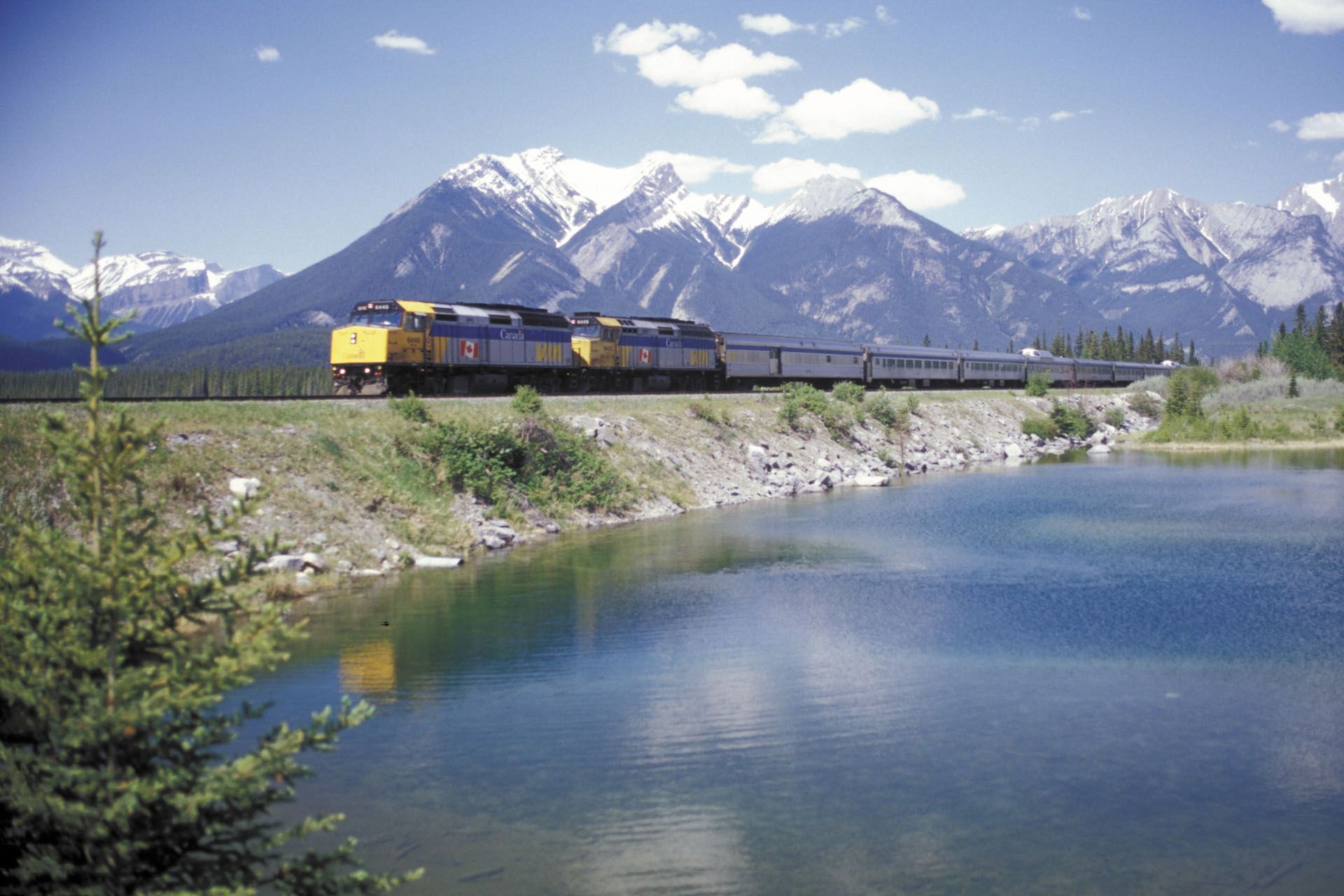 Train travels along tracks beside a lake with mountains in the background under a blue sky.