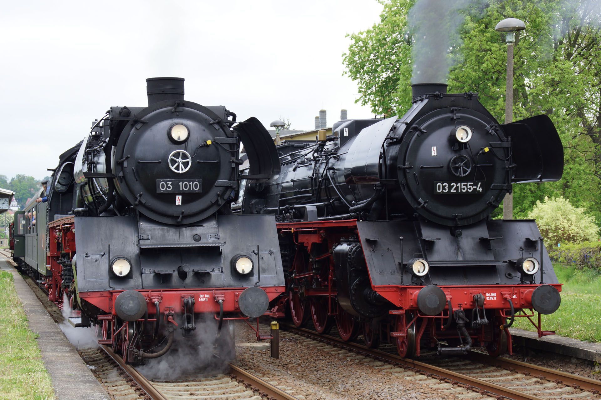 Two black steam locomotives side-by-side on tracks, with smoke billowing, red wheels, and facing the camera.