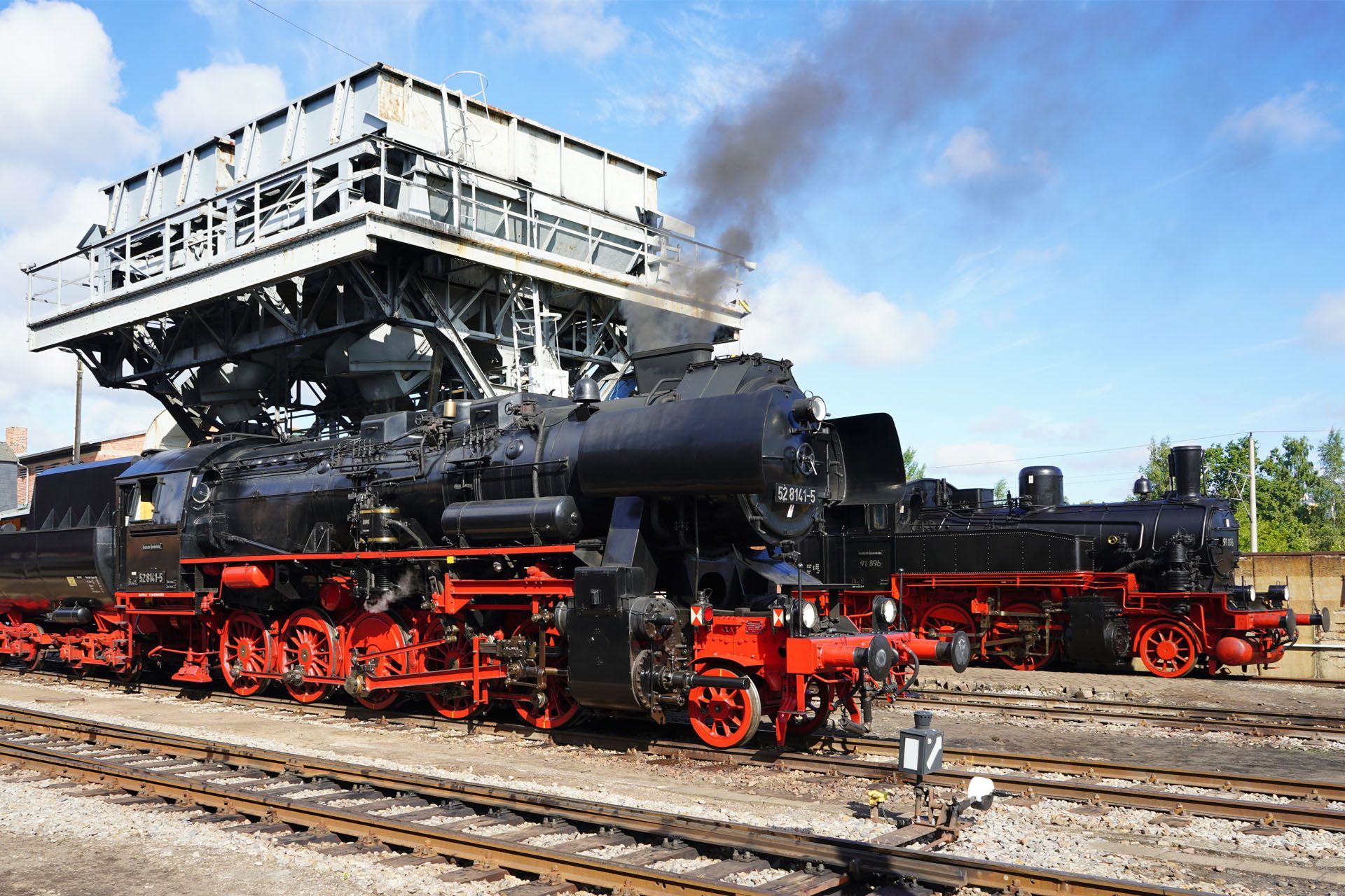 Two black and red steam locomotives on tracks, one under a coal loading structure.