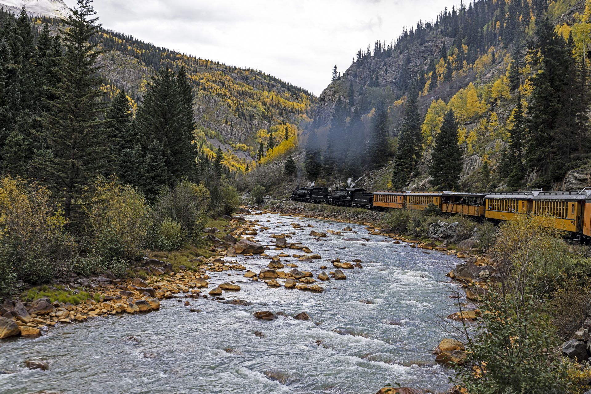 Steam train travels along a river through a valley with trees. Yellow train cars, autumn colors.