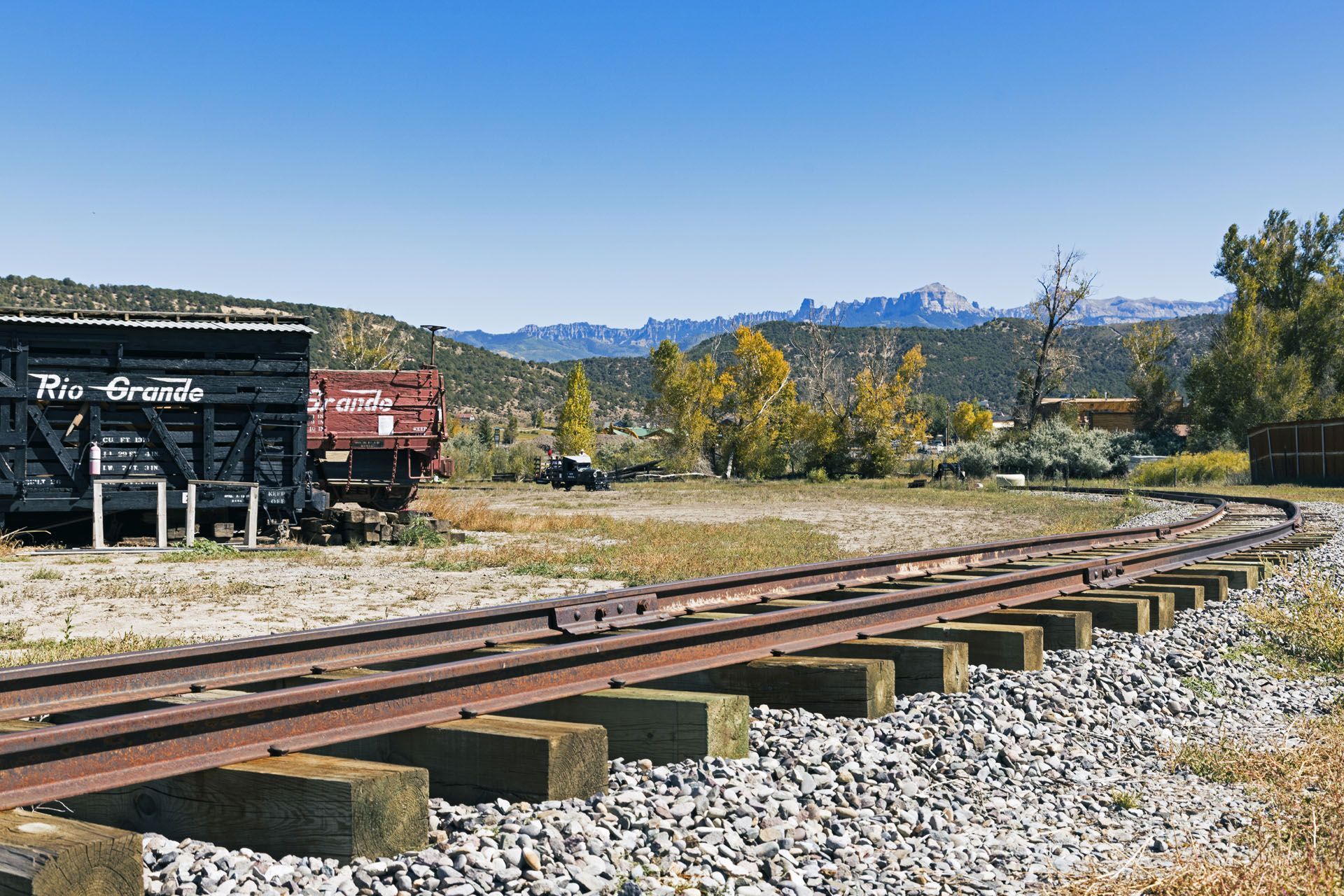 Railroad tracks curve past weathered train cars and a scenic landscape under a blue sky.