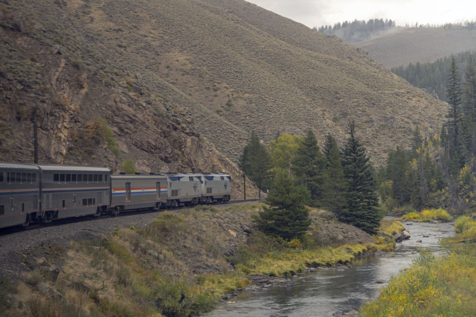 Train travels through a mountain canyon beside a river on a cloudy day.