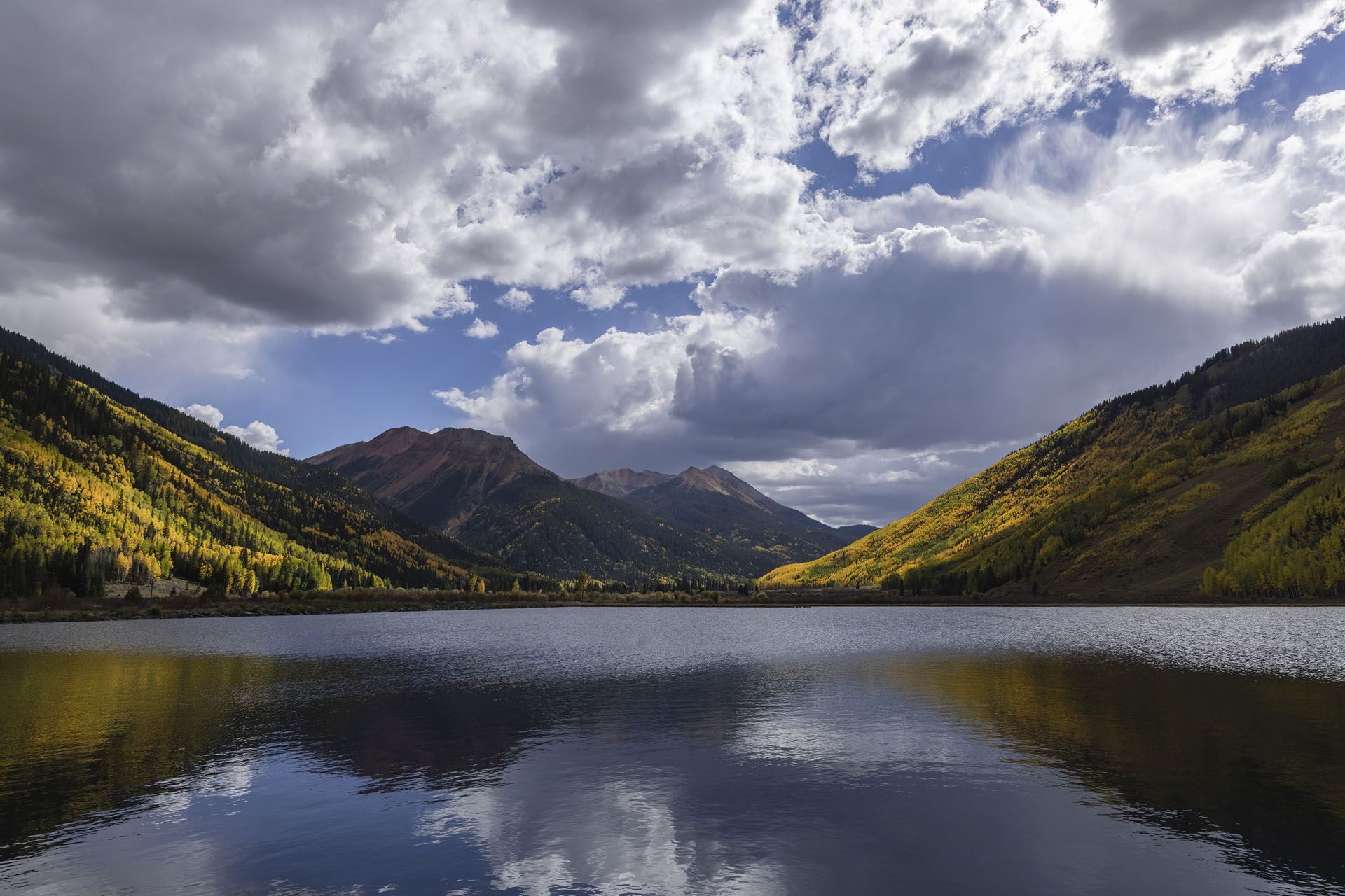 Lake surrounded by mountains and fall foliage under a cloudy sky reflecting in the water.