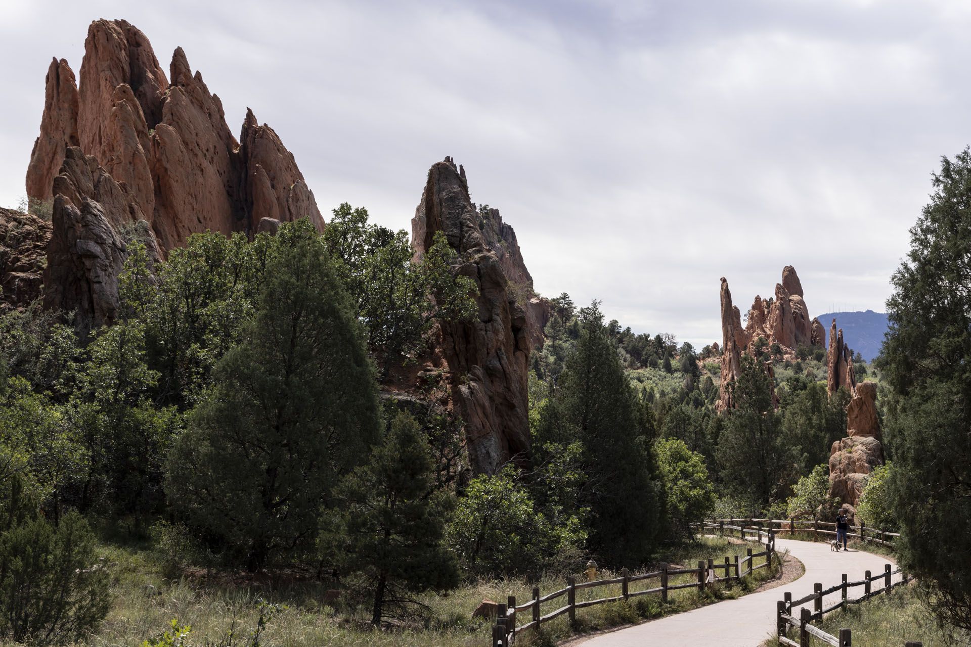 Red rock formations frame a winding path with a wooden fence. Lush green trees and vegetation dot the landscape under a cloudy sky.
