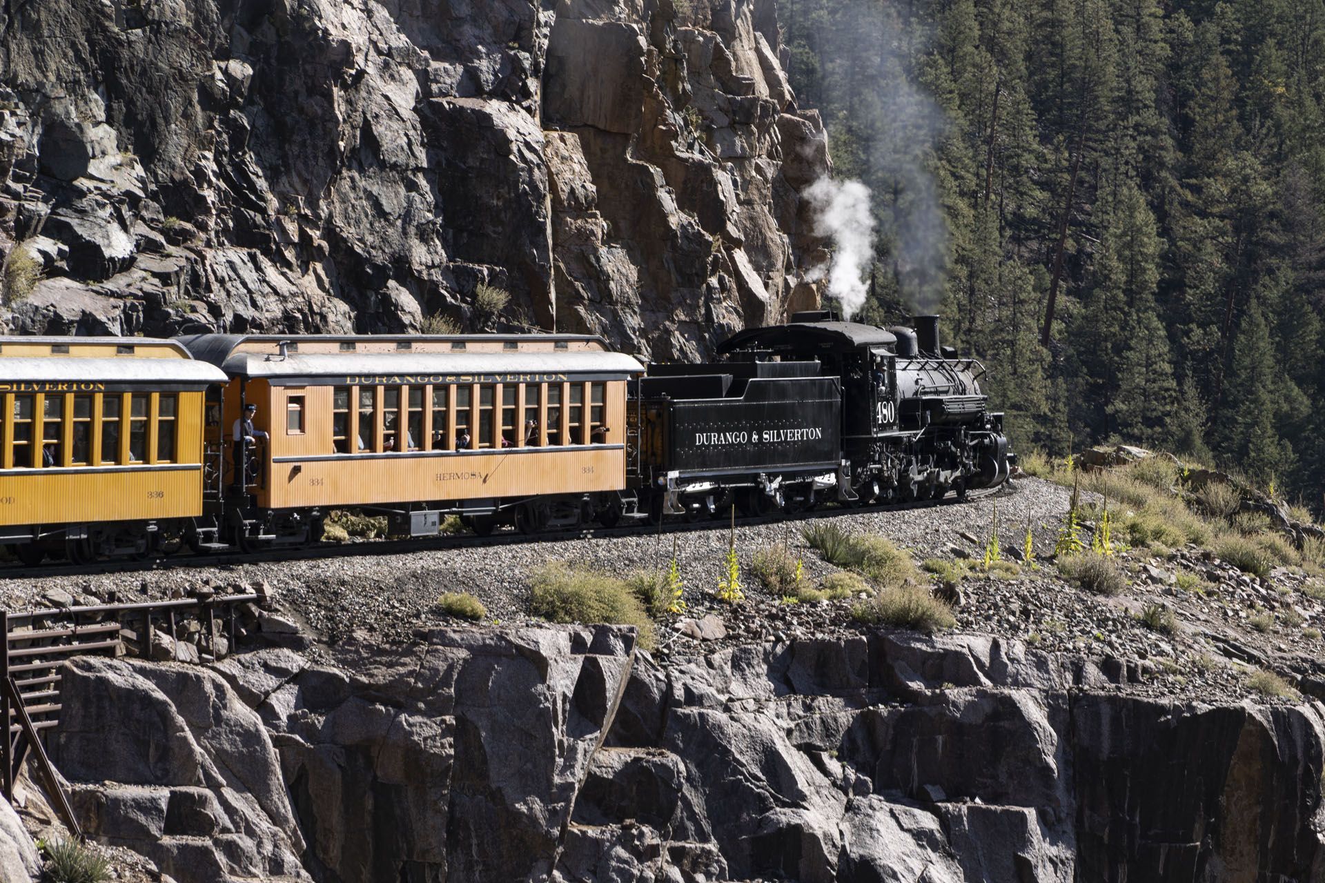 Steam train travels along a rocky mountain cliffside. Yellow passenger cars follow the black locomotive.