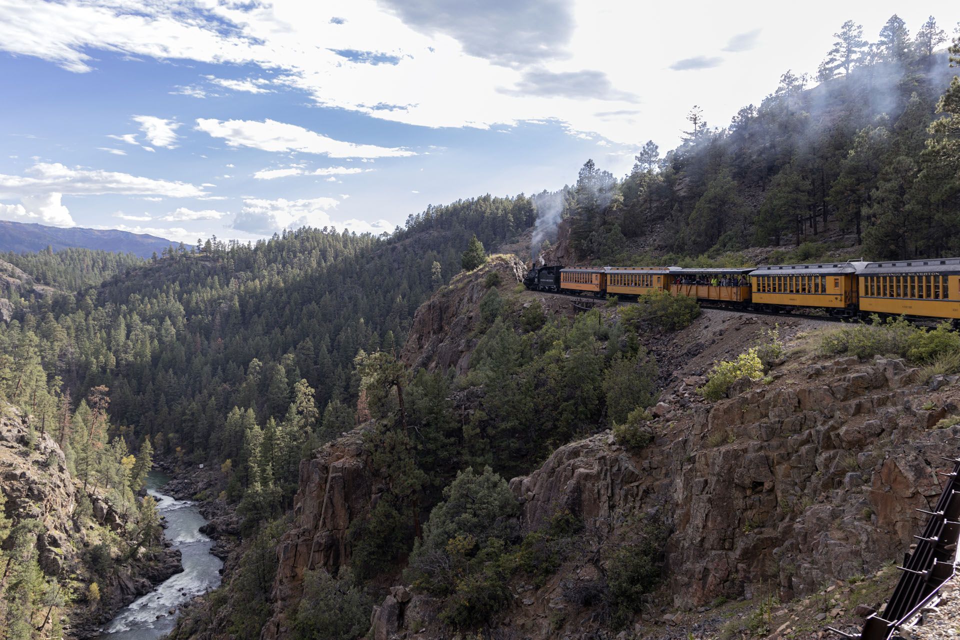 Steam train traveling along a cliffside beside a river through a mountain range.