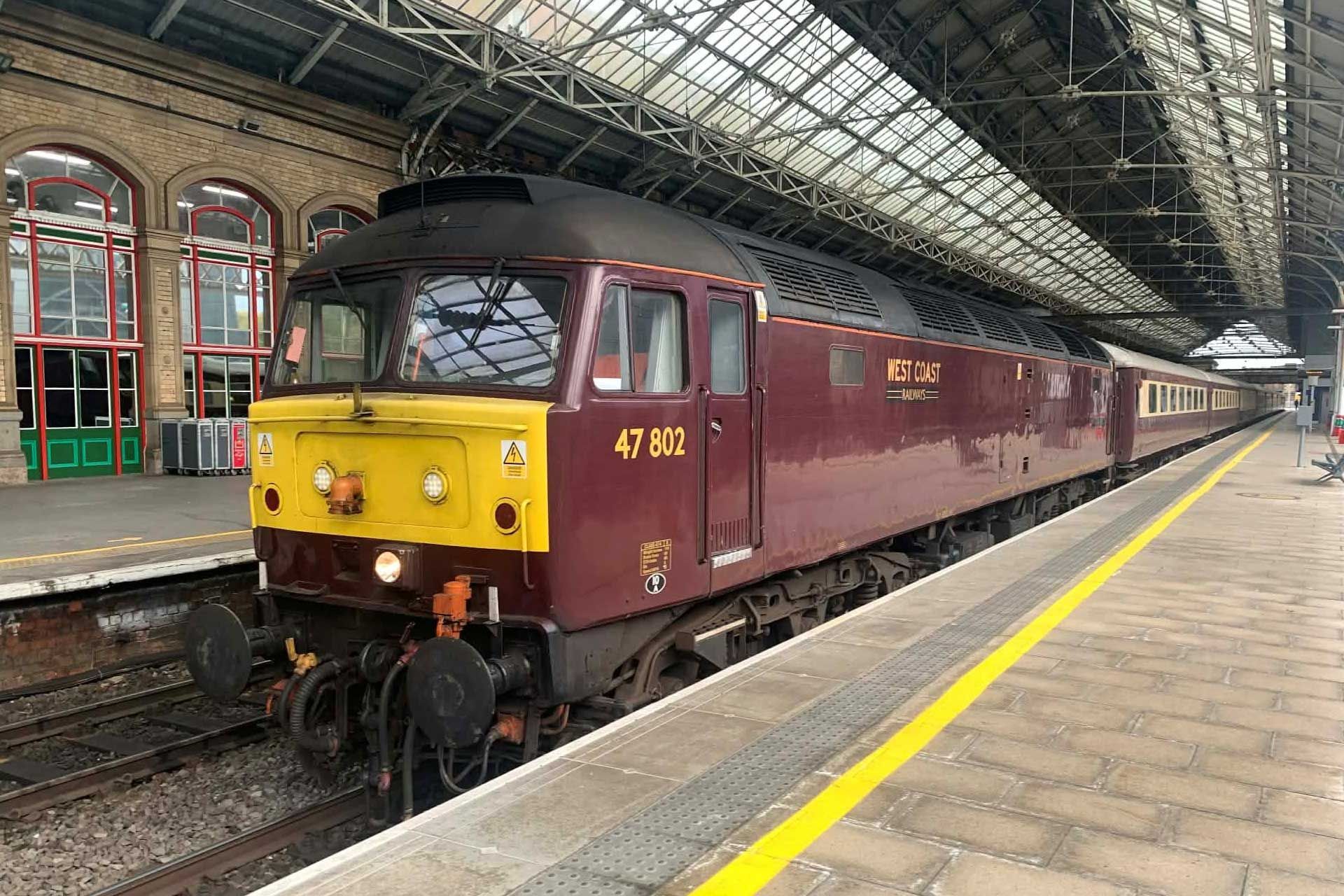 Burgundy train at station platform. Yellow front, dark red body. 47 802 on the side.