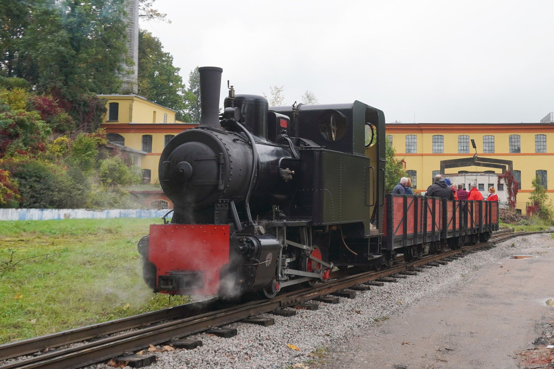 Black steam train pulling open red cars along tracks, with passengers in the cars.