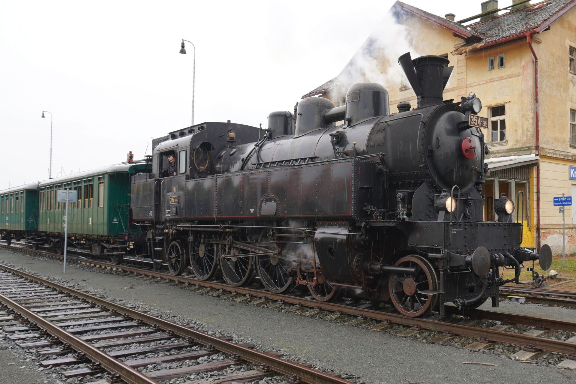 Black steam train with green passenger cars on tracks next to a station building.