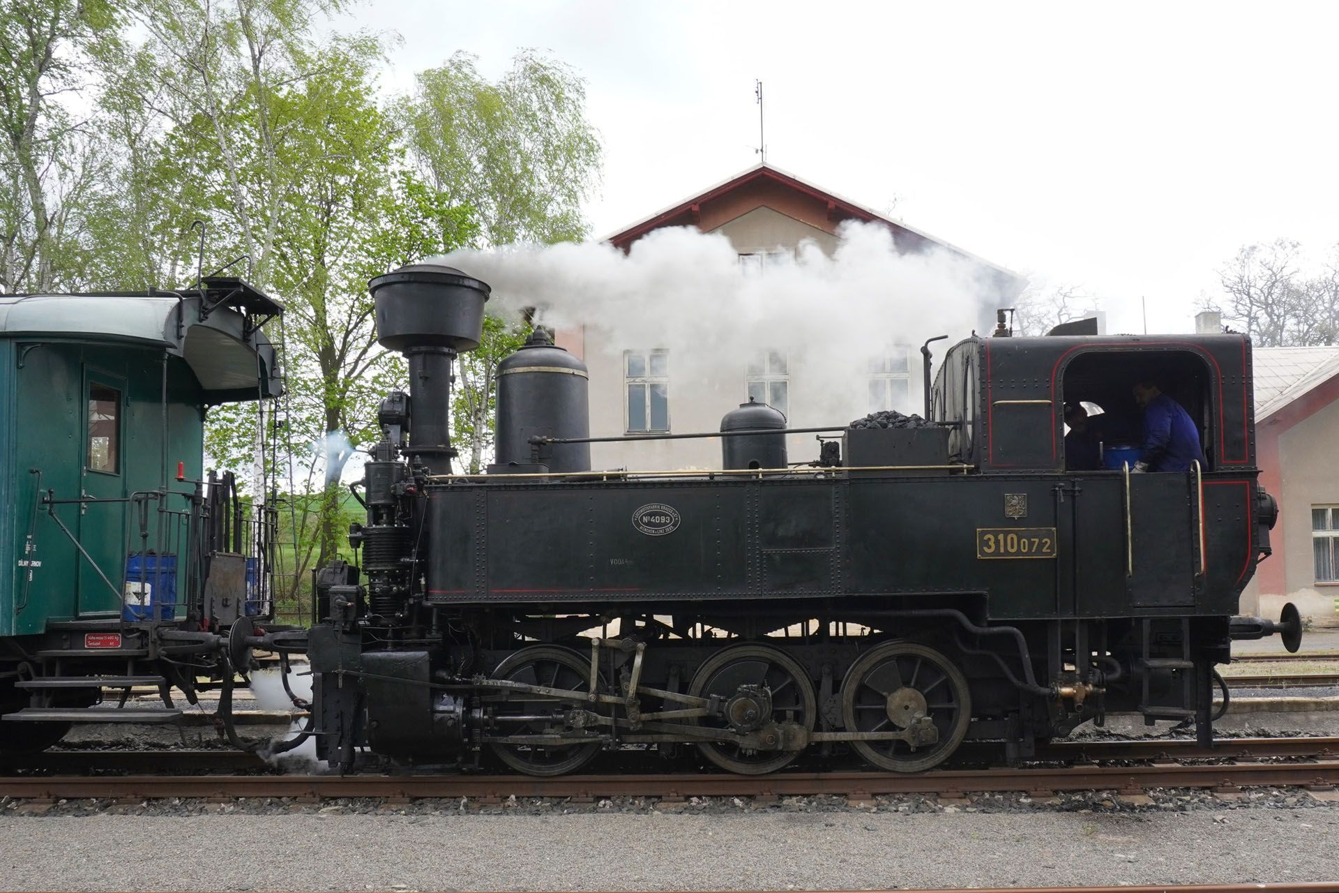 Steam train next to a green train car, emitting steam. Building in the background.