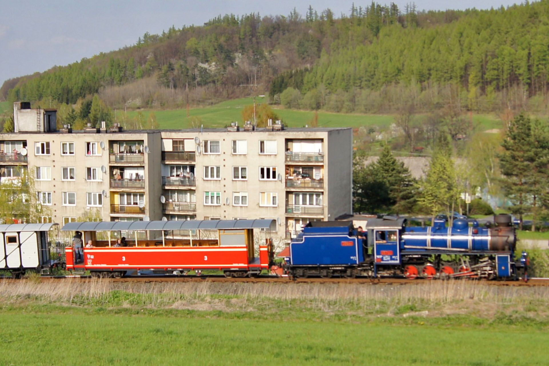 A blue steam locomotive pulls an open-air red passenger car past an apartment building near a grassy hill.