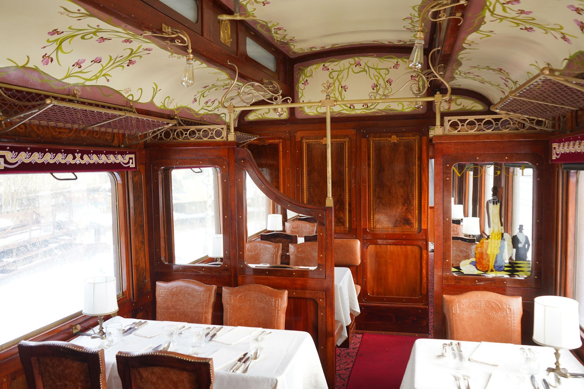 The interior of a vintage train dining car with polished wooden paneling, white-clothed tables, and floral ceiling art.