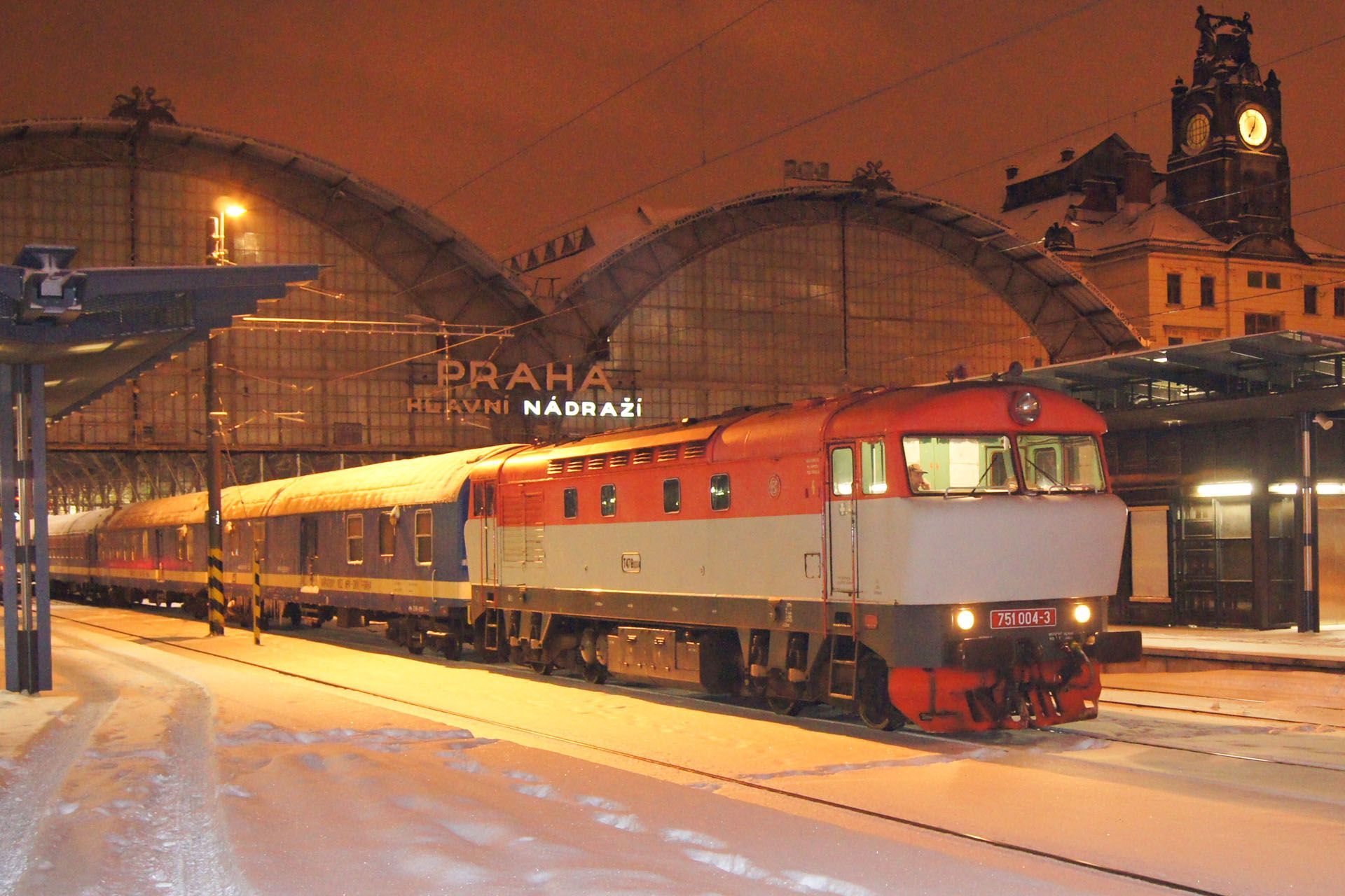 A diesel train sits on snowy tracks at night in front of the illuminated Prague Main Station building.