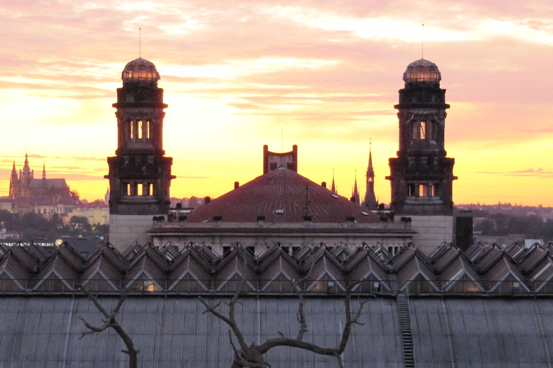 Prague Main Railway Station's Art Nouveau domes backlit by a sunset, with St. Vitus Cathedral visible in the distance.