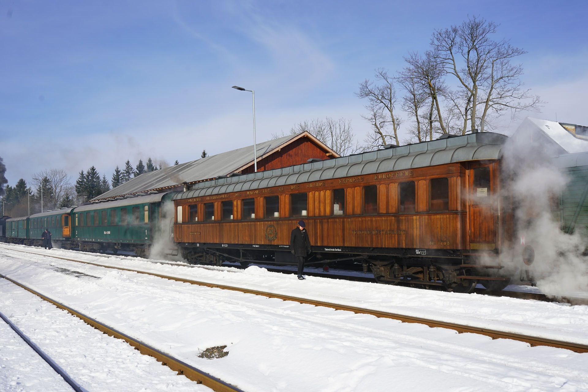 A wooden train carriage and green passenger cars stand at a snowy station platform on a sunny day.