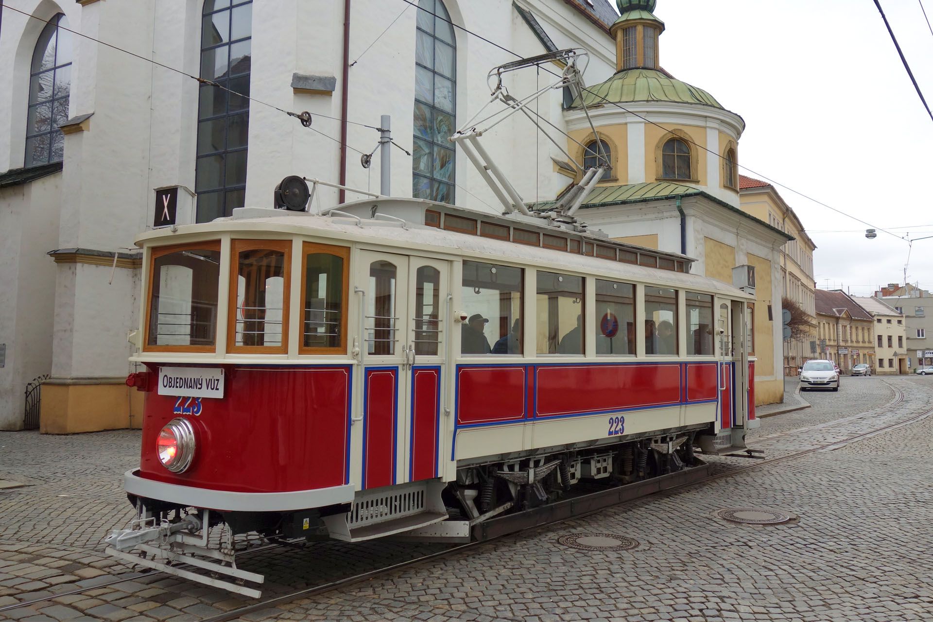 A vintage red and cream streetcar on cobblestone tracks in front of a white church in a European-style town.
