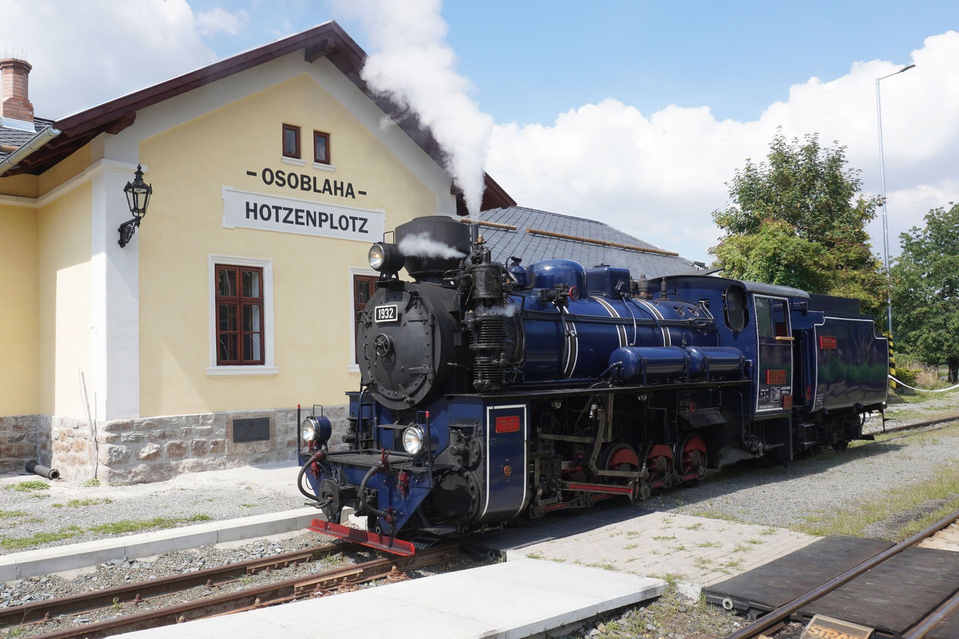 A blue steam locomotive emits white smoke while stopped at a yellow train station building labeled 