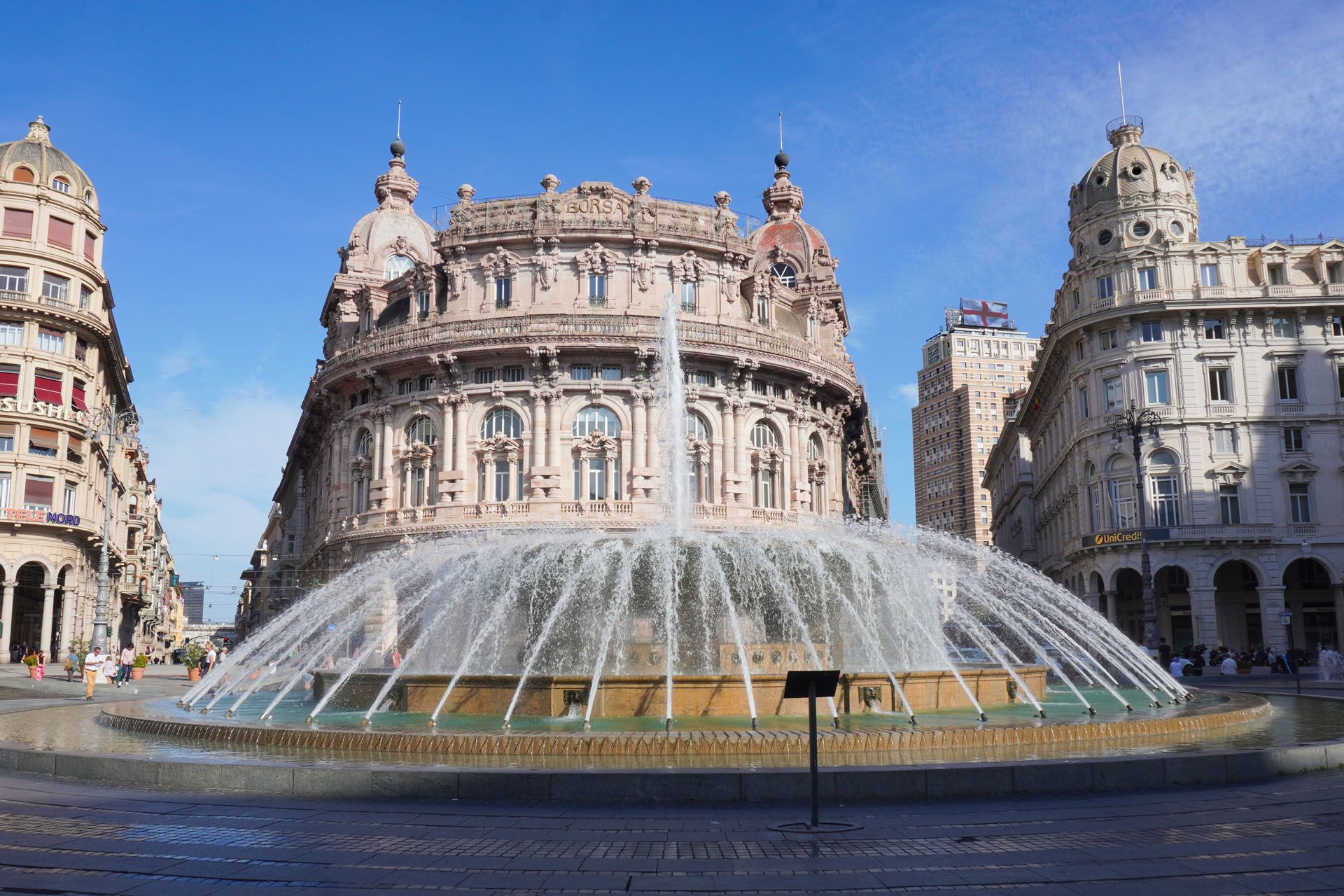 Large fountain in front of ornate building. Water arcs into a circular pool. Buildings line the square in Genoa, Italy.