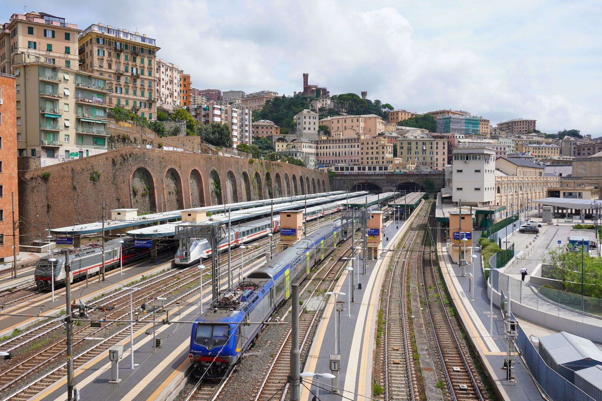 Railway station with multiple tracks, trains, and surrounding buildings in Genoa, Italy.
