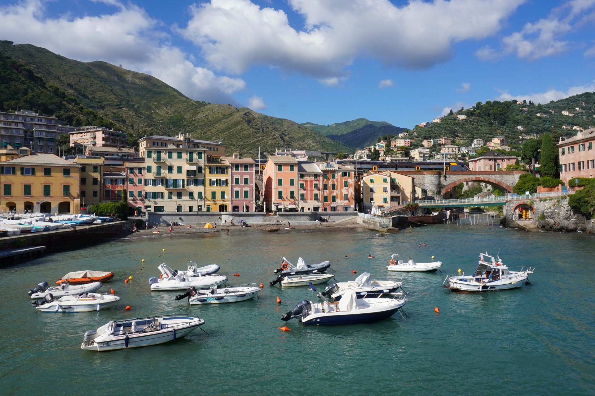 Boats float in a harbor with colorful buildings, bridge, and mountains in the background. Blue water and sky.