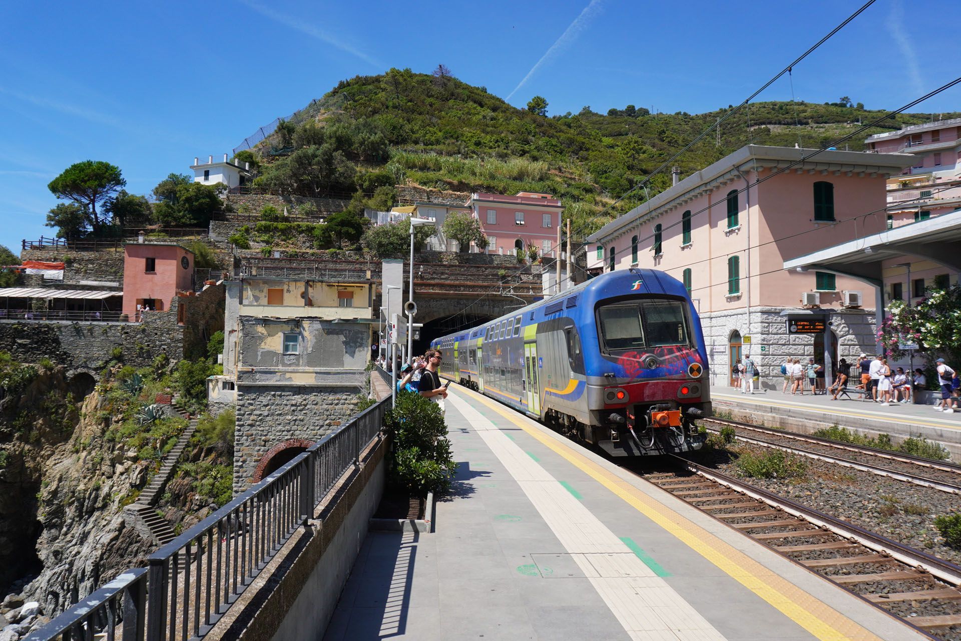 Train pulling into a station in a mountainous coastal town under a blue sky.