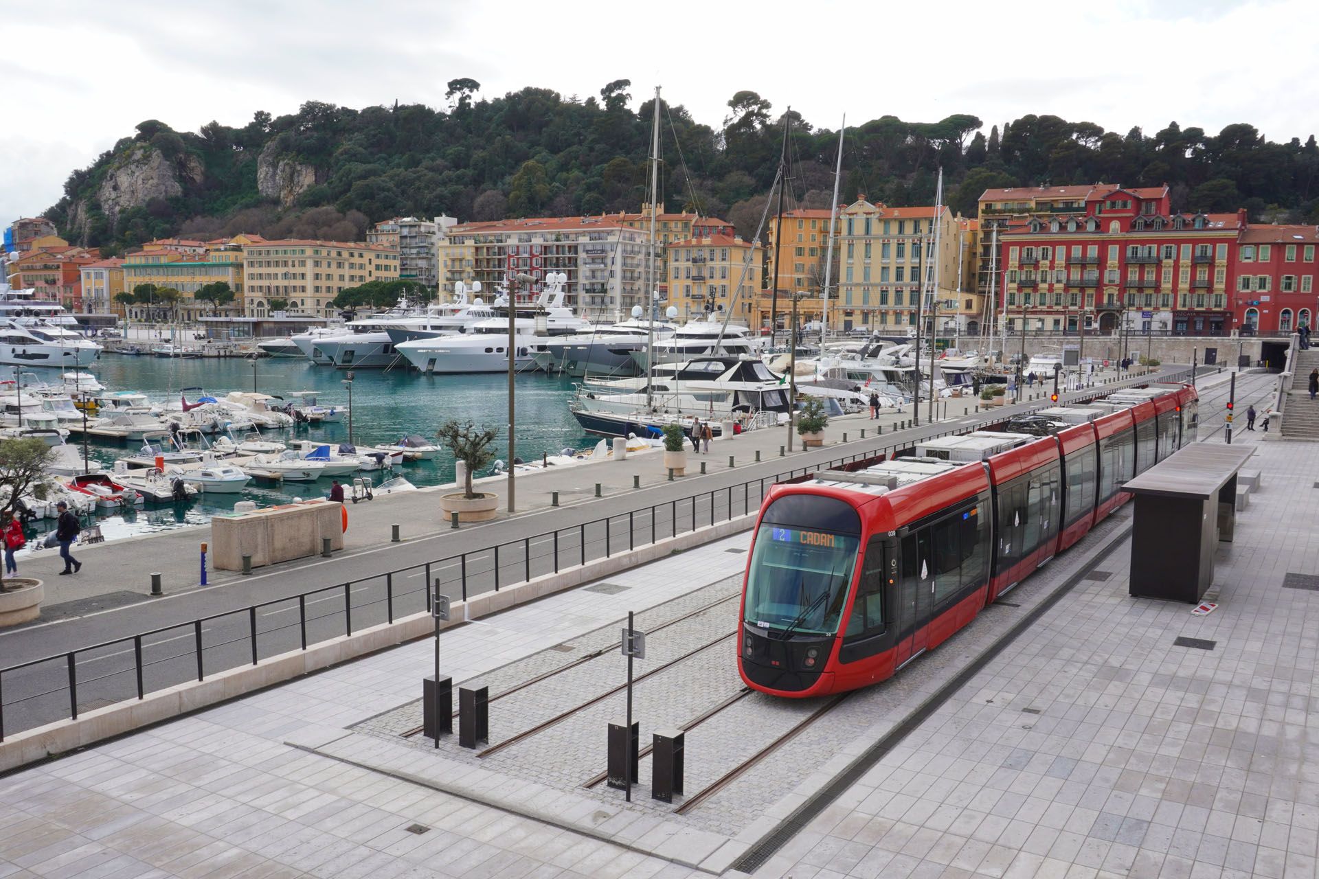 Red tram on tracks in a harbor, boats docked, colorful buildings in background, hillside.