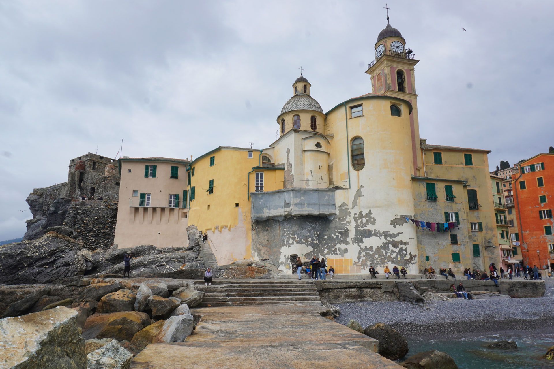 Church by the sea with a clock tower and dome, people walking on stone walkway, overcast sky.