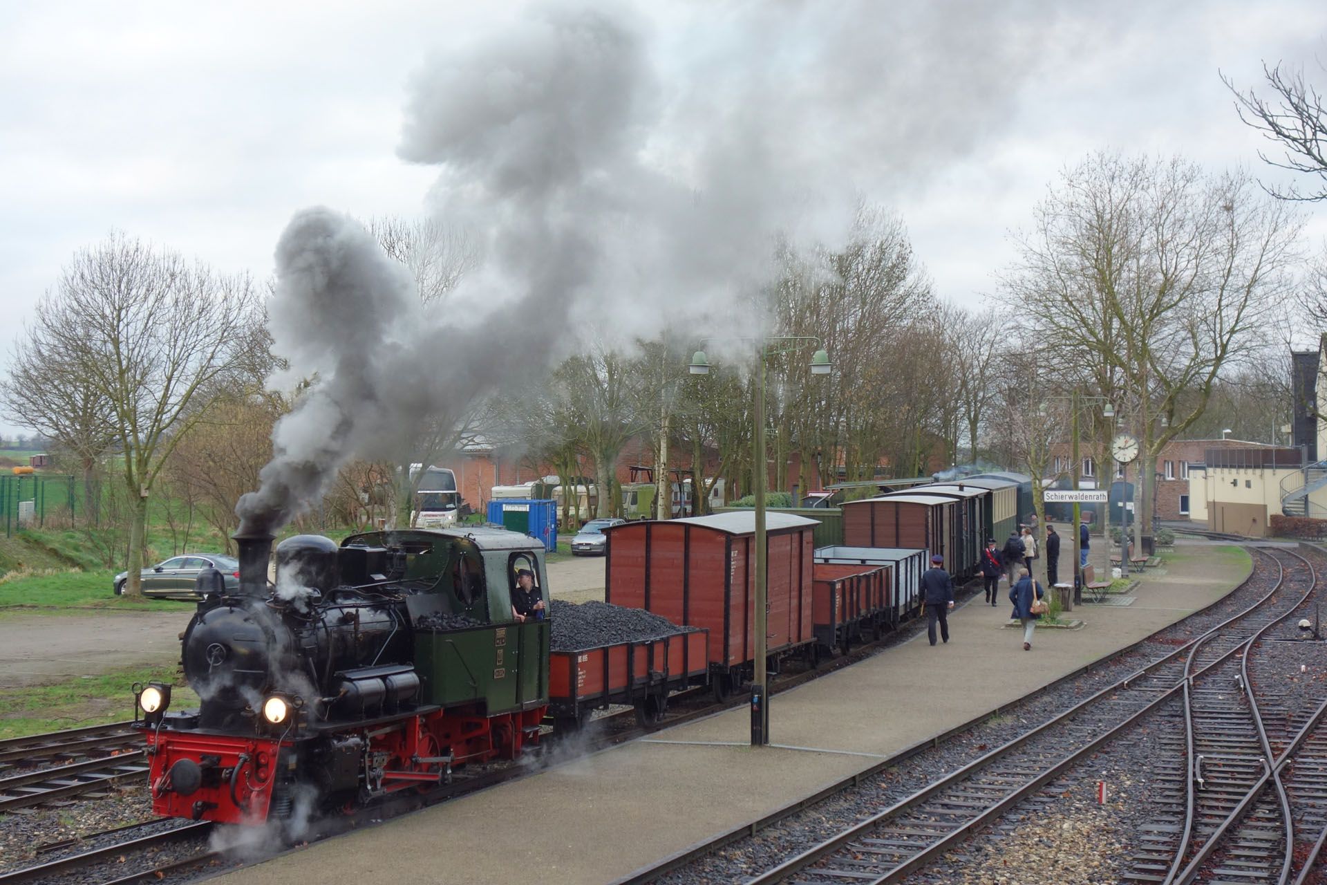 Steam train at a station, billowing smoke. Red, green, and black engine pulling brown freight cars.