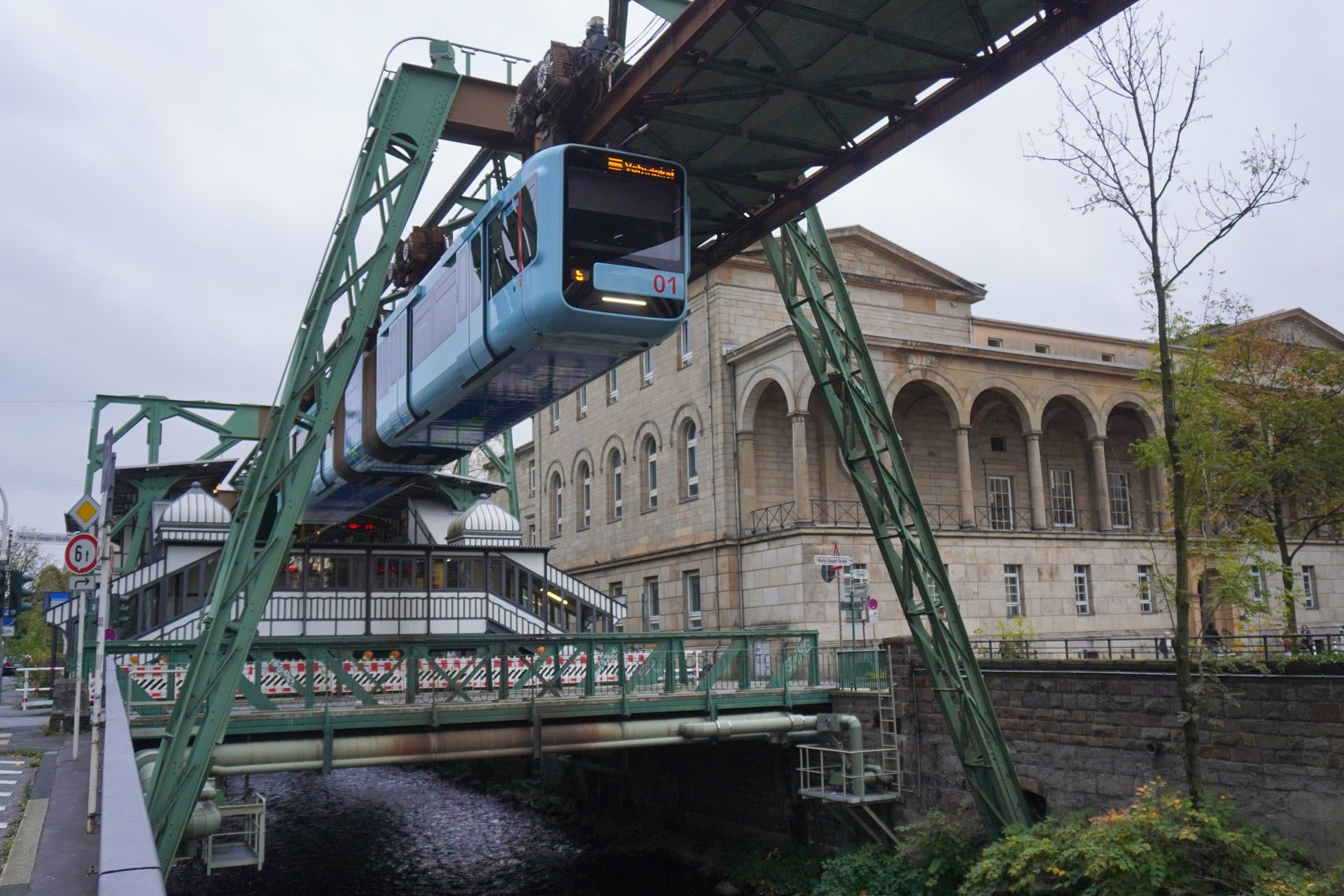 Blue suspension train traveling over a river, with a historic building in the background. Green support structures and overcast sky.