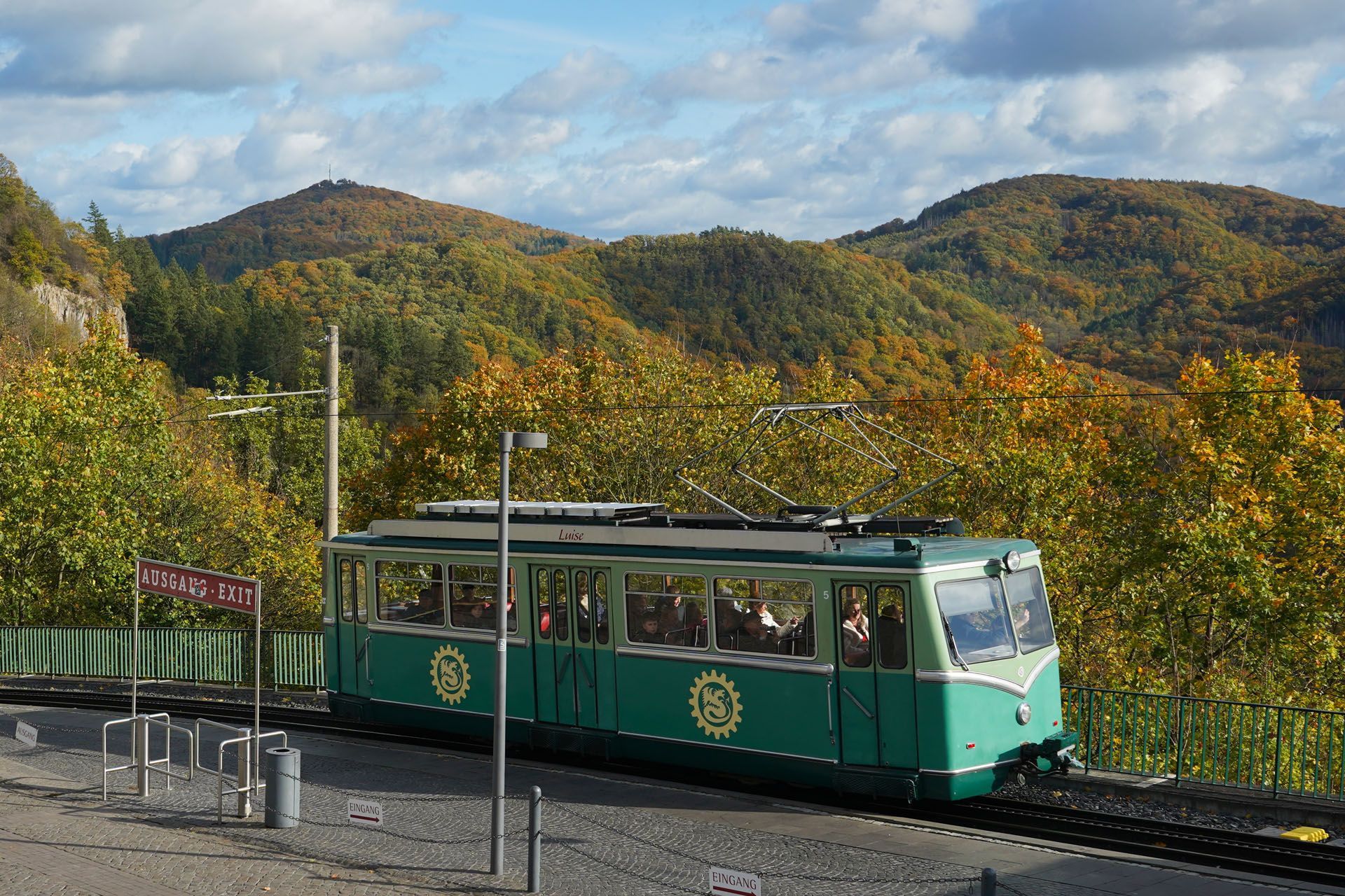 Green train on tracks near mountains with autumn foliage.