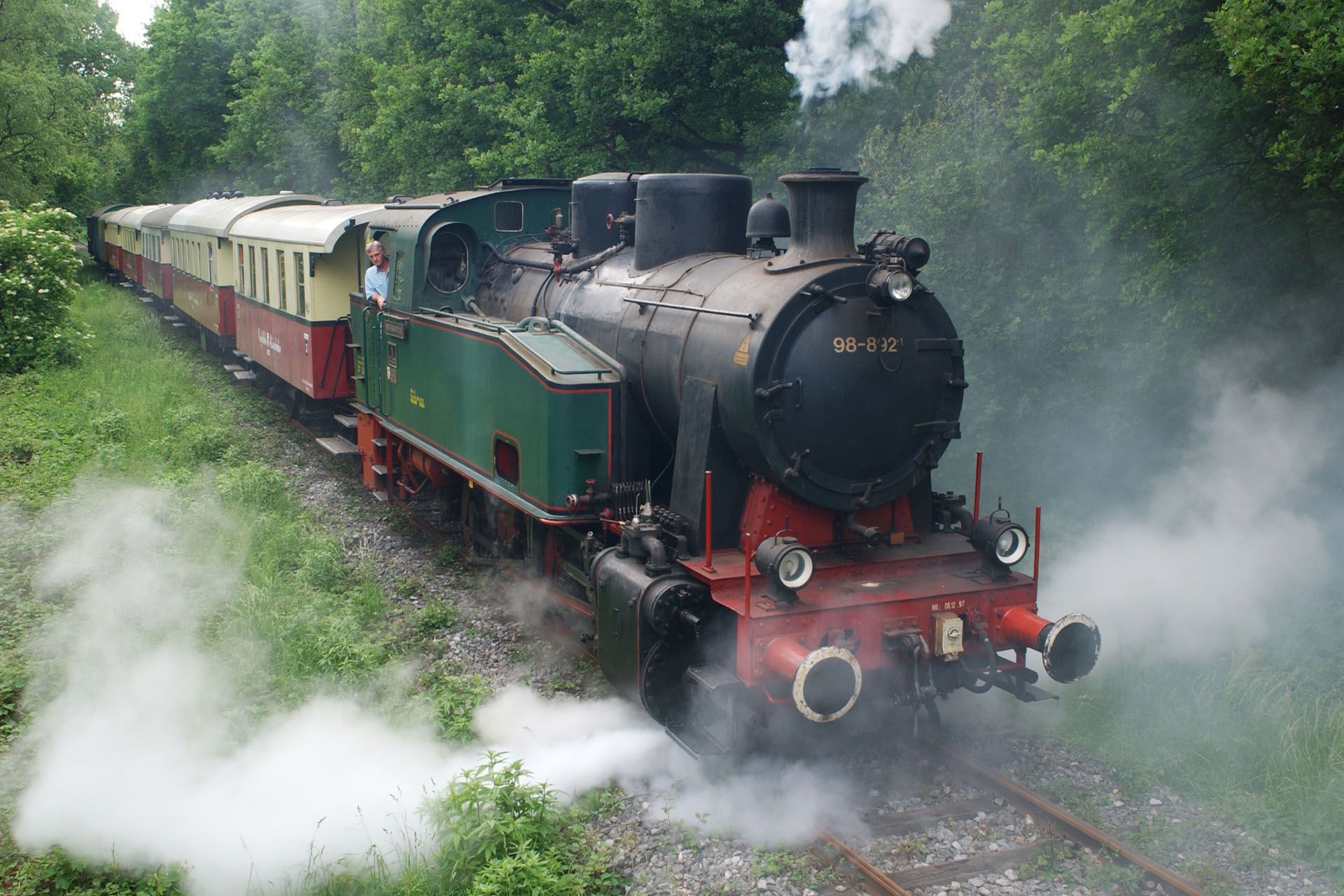 Steam train on tracks surrounded by greenery. Black engine, red and cream carriages, white steam.