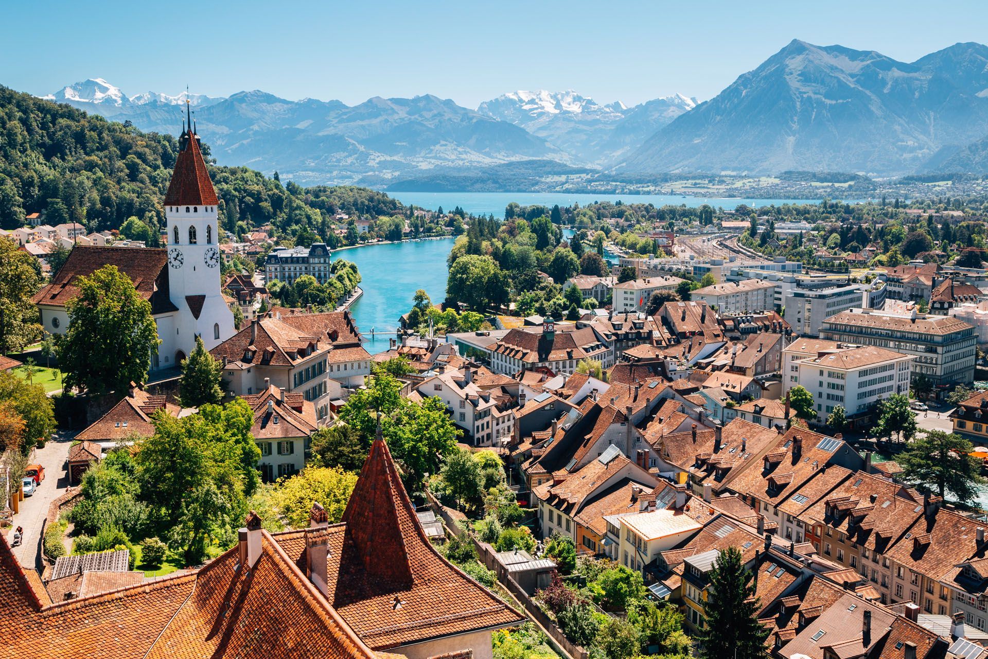 Cityscape of Thun, Switzerland, with red-roofed buildings, lake, mountains, and a white church tower.