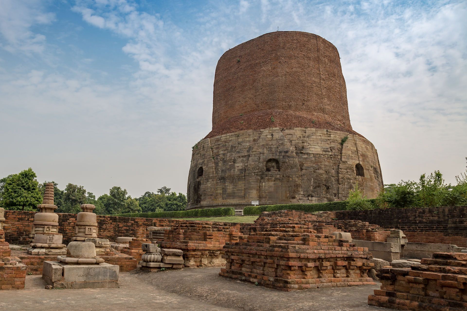 Ancient brick stupa ruins in a courtyard with scattered stone structures and trees under a cloudy sky
