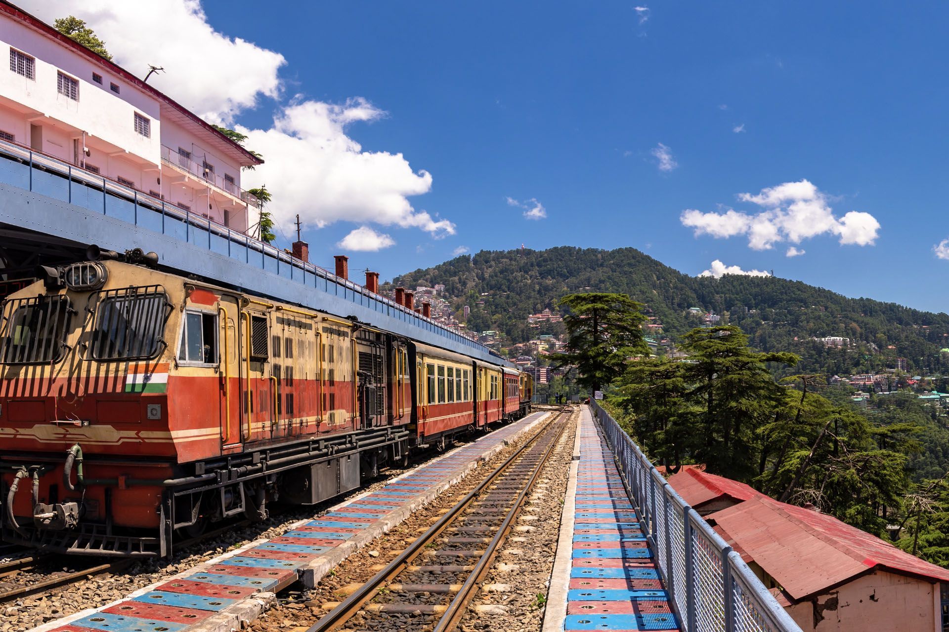 Red train on a mountain bridge beside blue tracks, with hills and a cloudy sky in the background