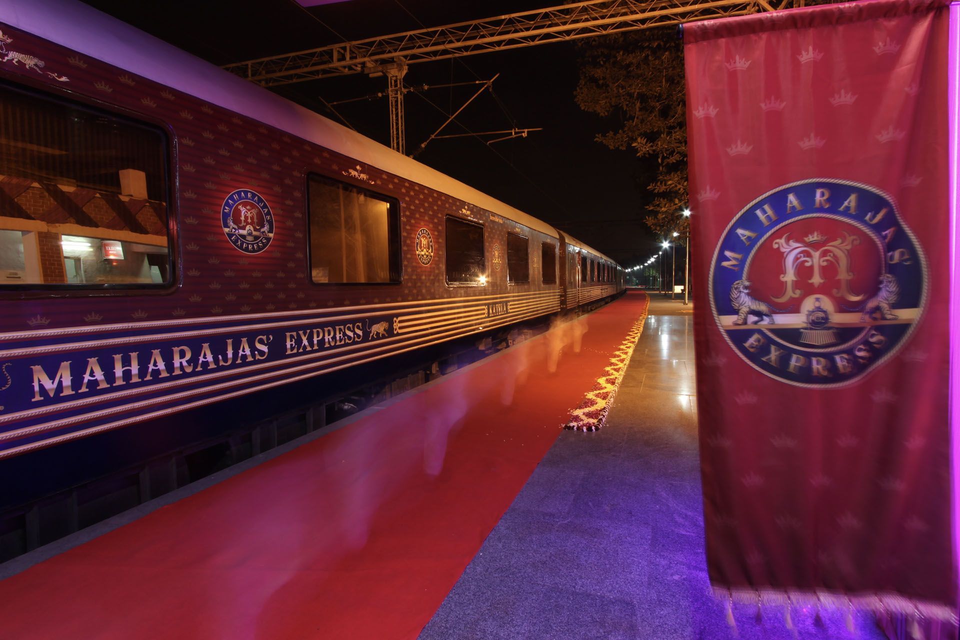Purple-lit Maharajas Express train at night, with a red carpet and branded banner beside it