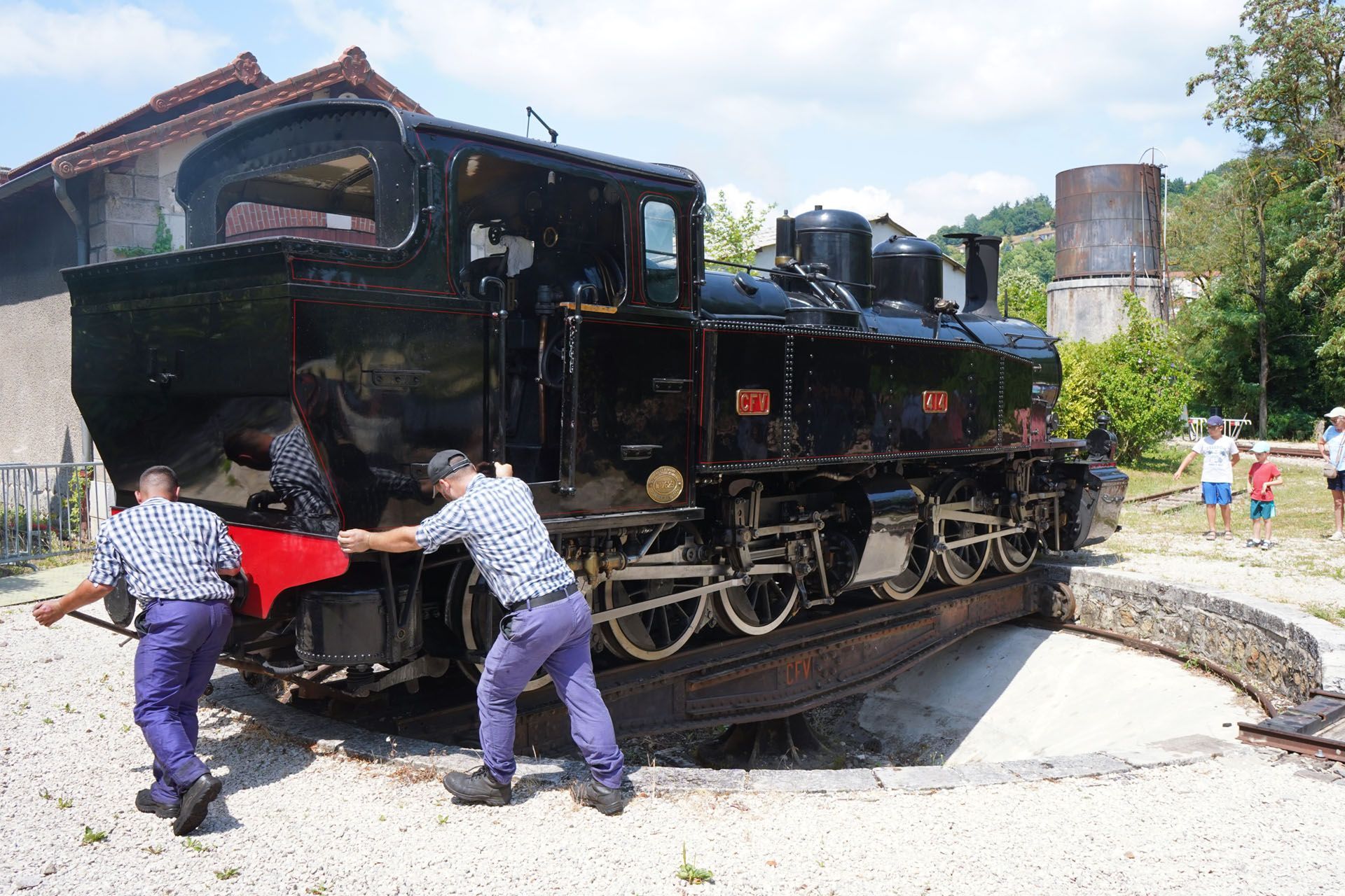 Two men turning a black steam engine on a circular track. Sunny day.