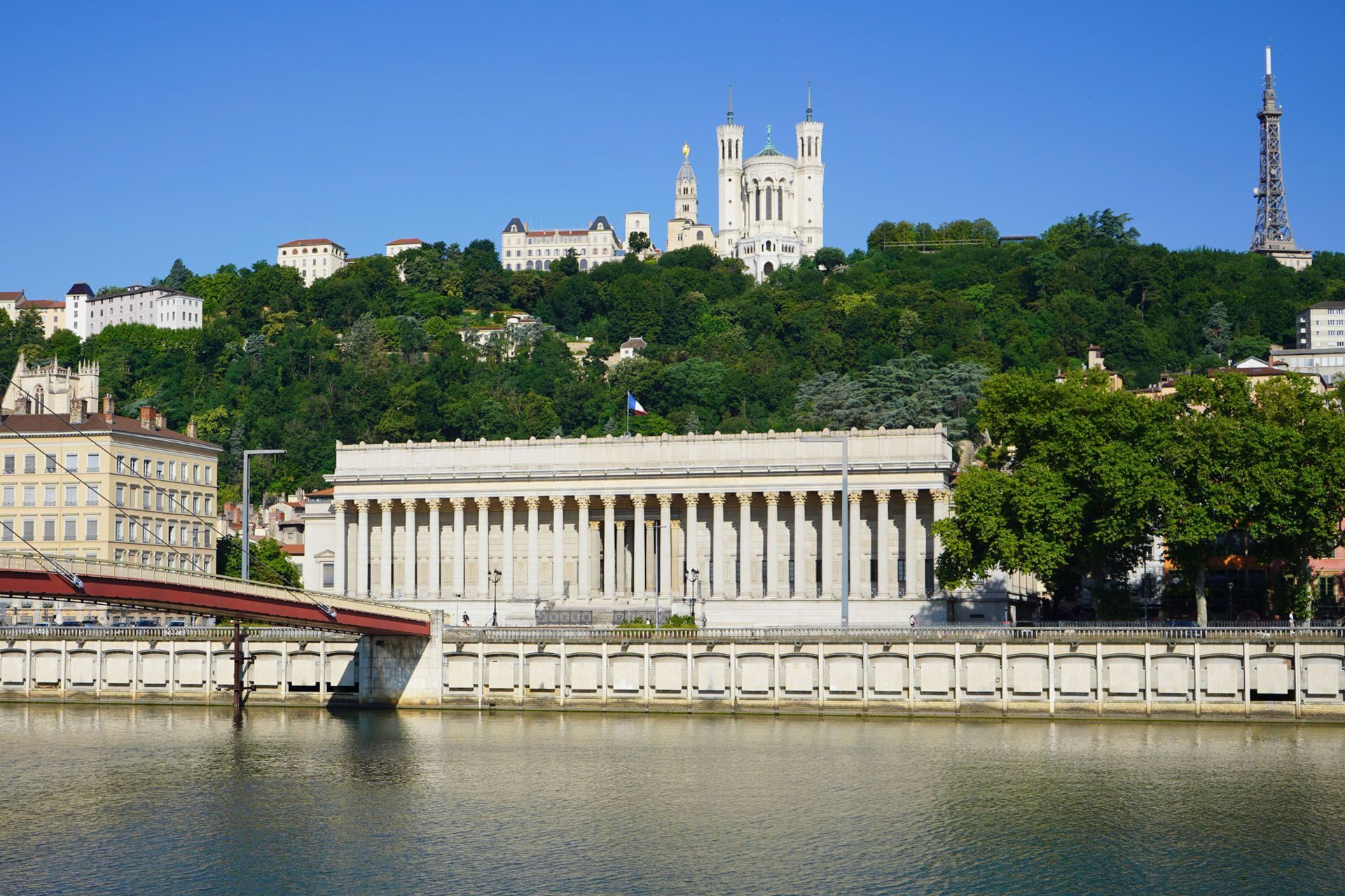 Classical building and bridge on a river, with a church and tower on a green hill, under a clear blue sky.