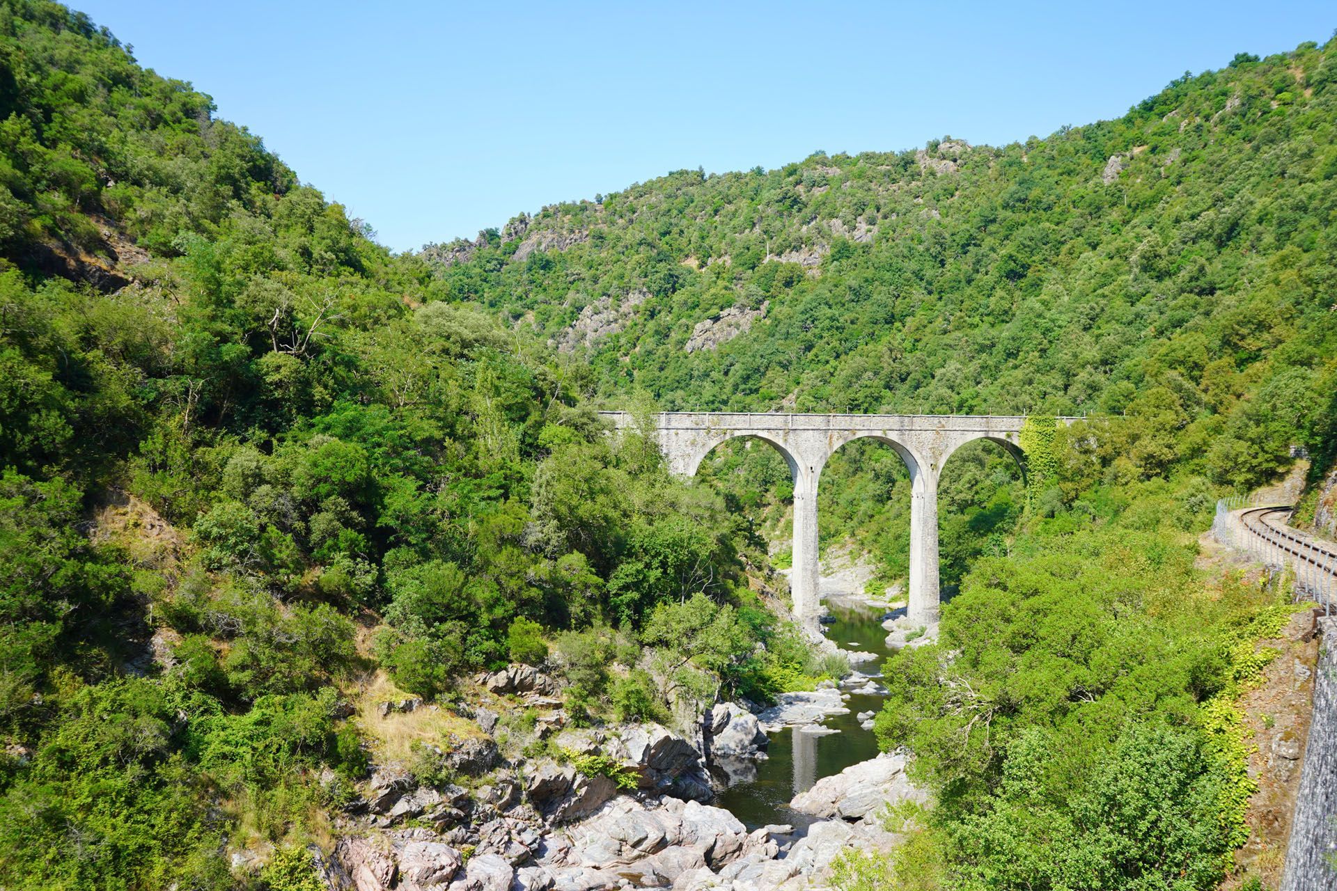 Stone arched bridge over a river, surrounded by green trees on a sunny day.