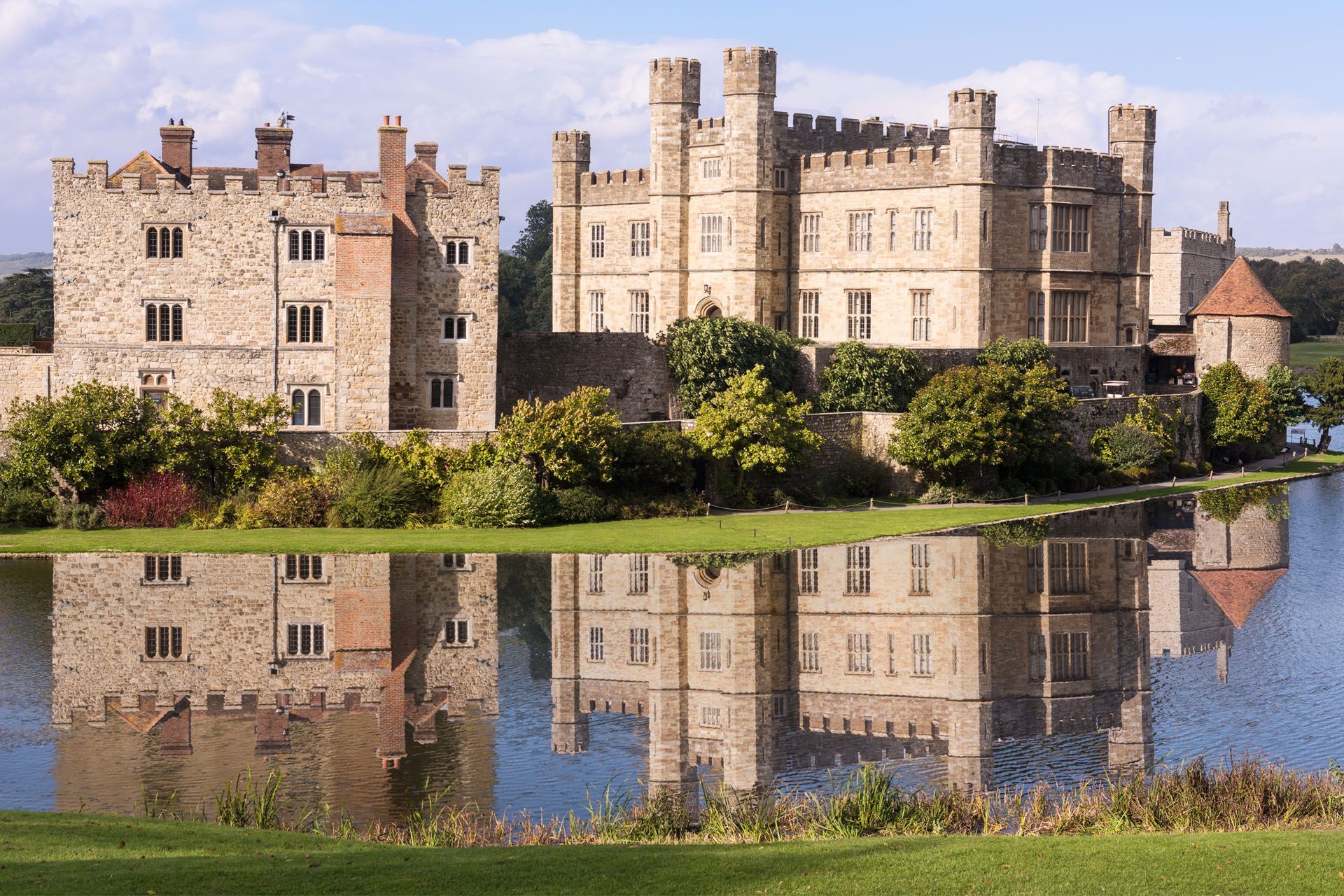 Leeds Castle reflected in water on a sunny day. Light stone castle, lush green grass, blue sky.