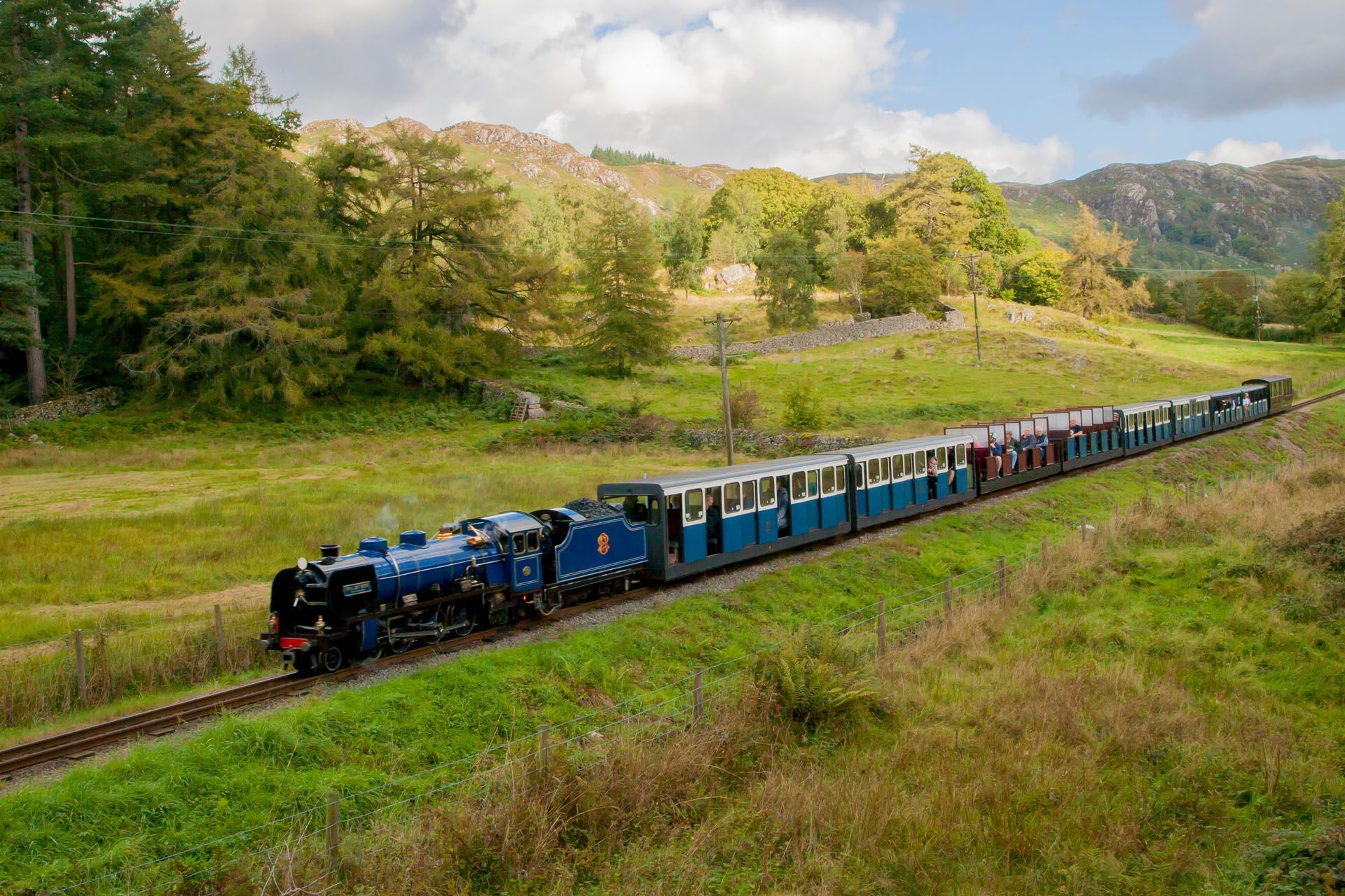 Blue steam train travels along tracks through a green field, with hills and trees in the background.