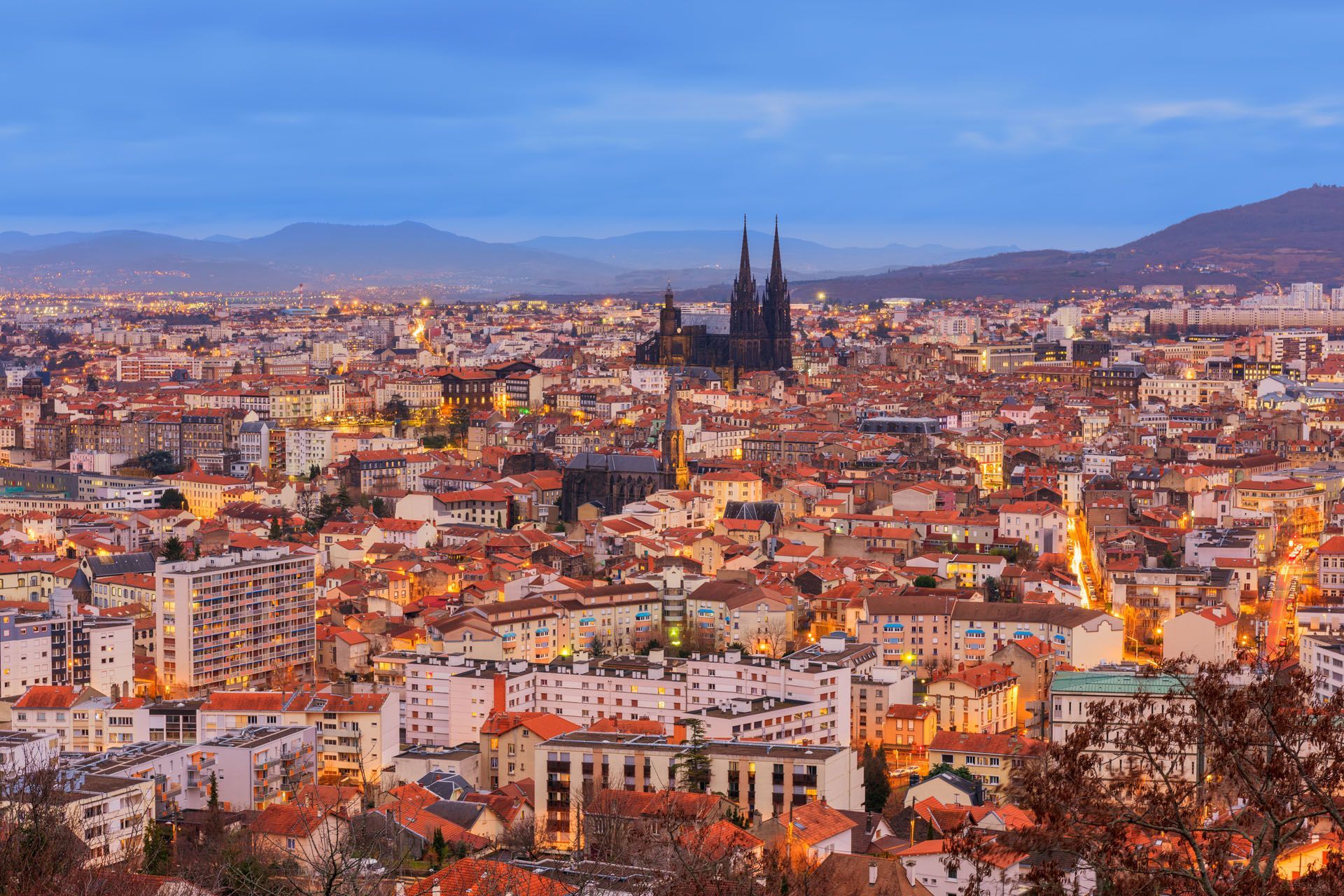 Cityscape at dusk, orange rooftops, cathedral spires rise above buildings, mountains in background.