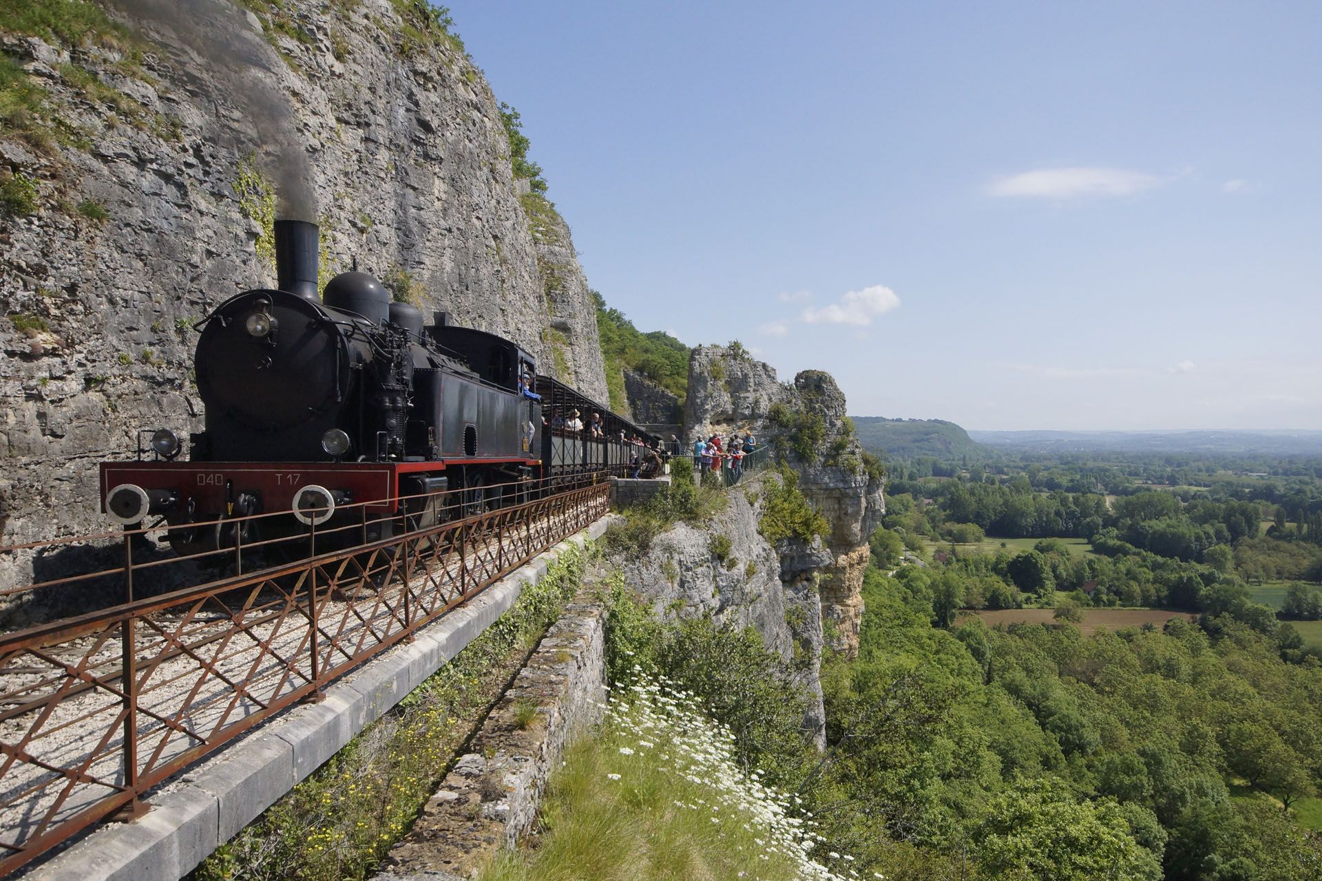 Steam train travels along a cliffside track, overlooking a green valley.