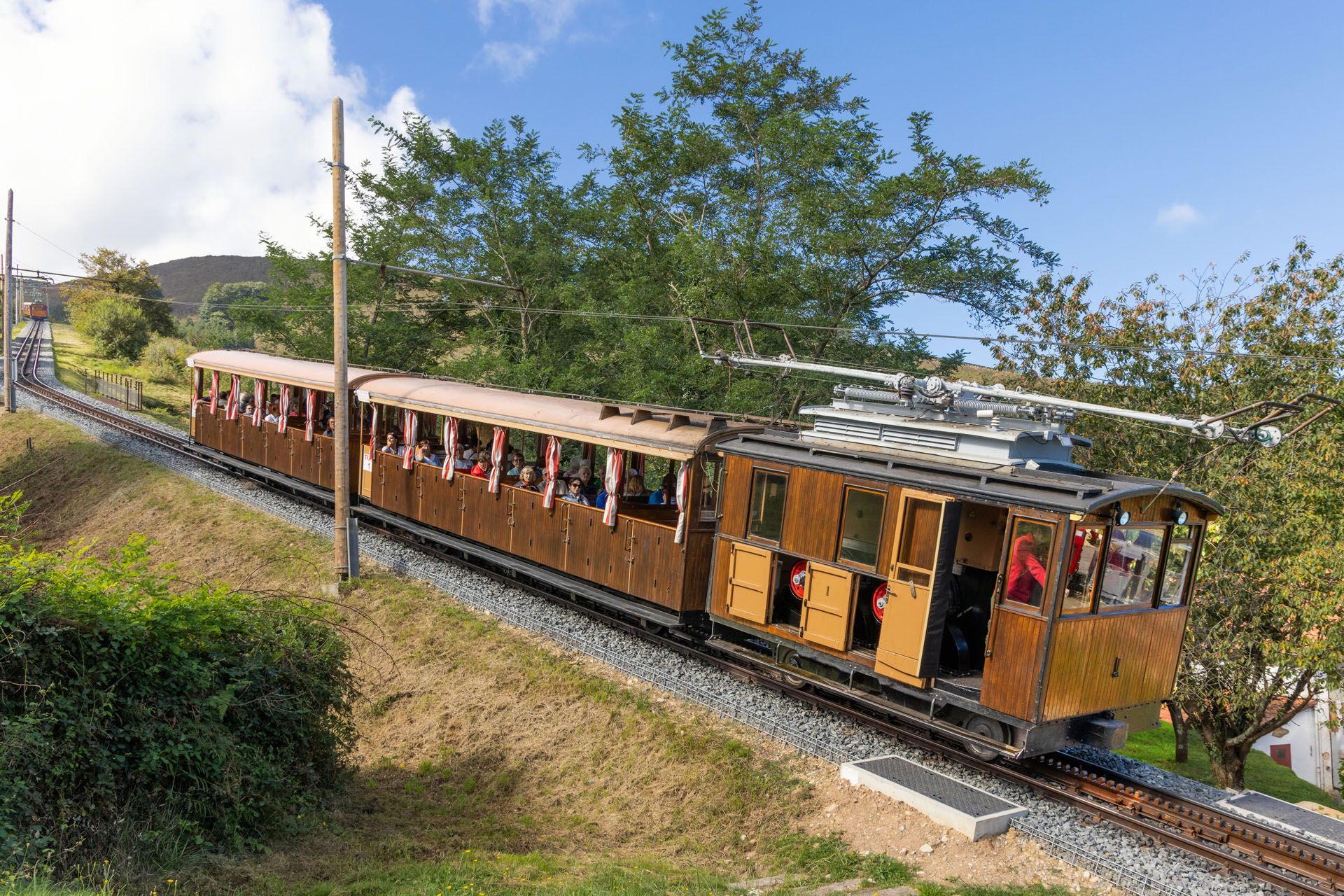 Wooden funicular train ascending a hillside on a sunny day; passengers visible inside.