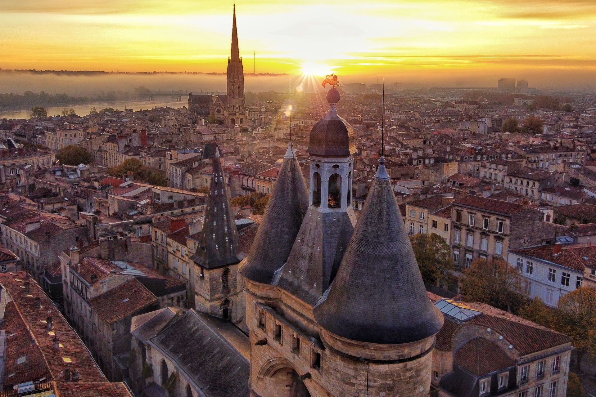 Overhead view of a European city at sunset, with a prominent stone gate and spire. Orange and gold hues dominate.