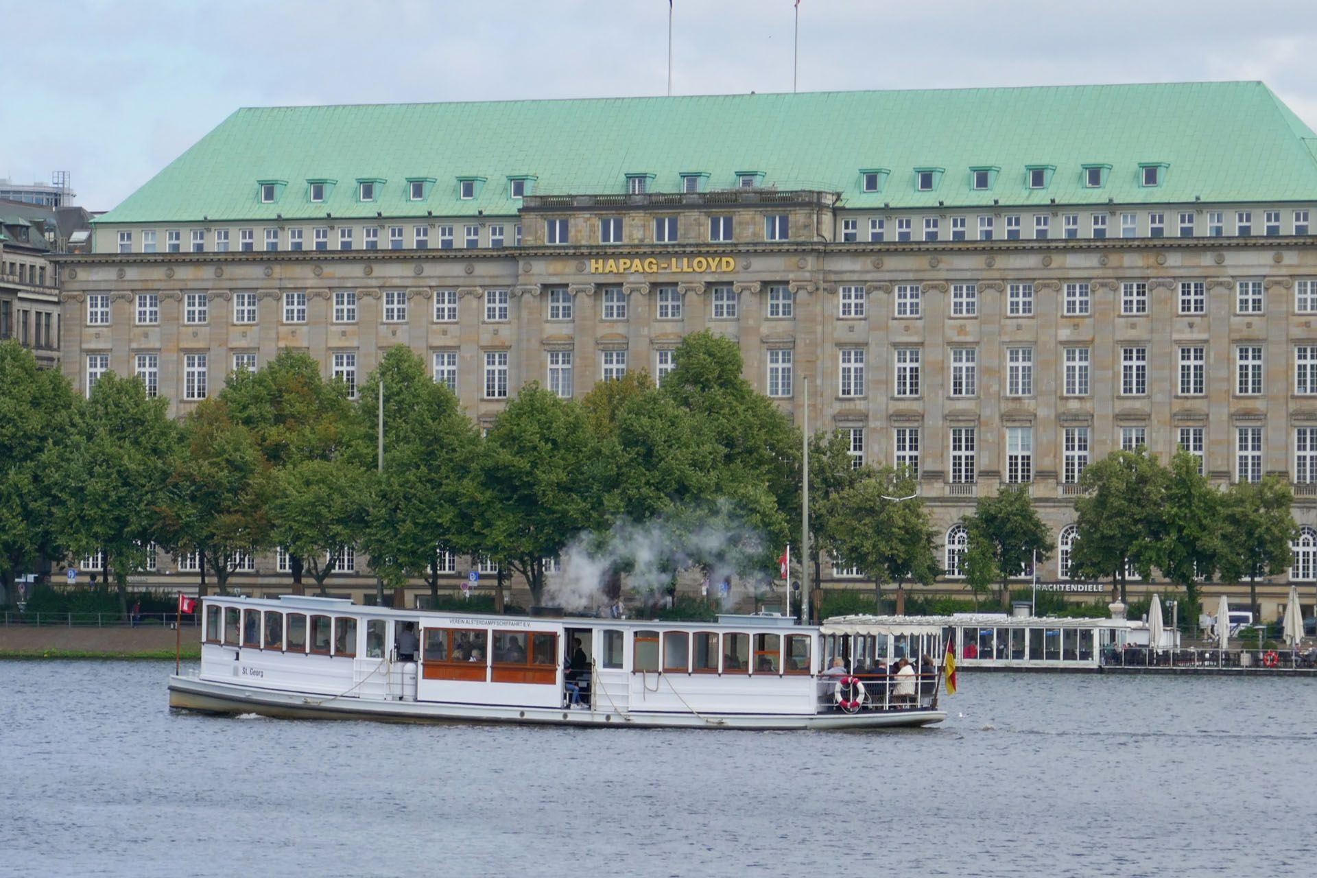Steamboat on a lake, smoke rising. Large building with green roof in the background. Hamburg, Germany.