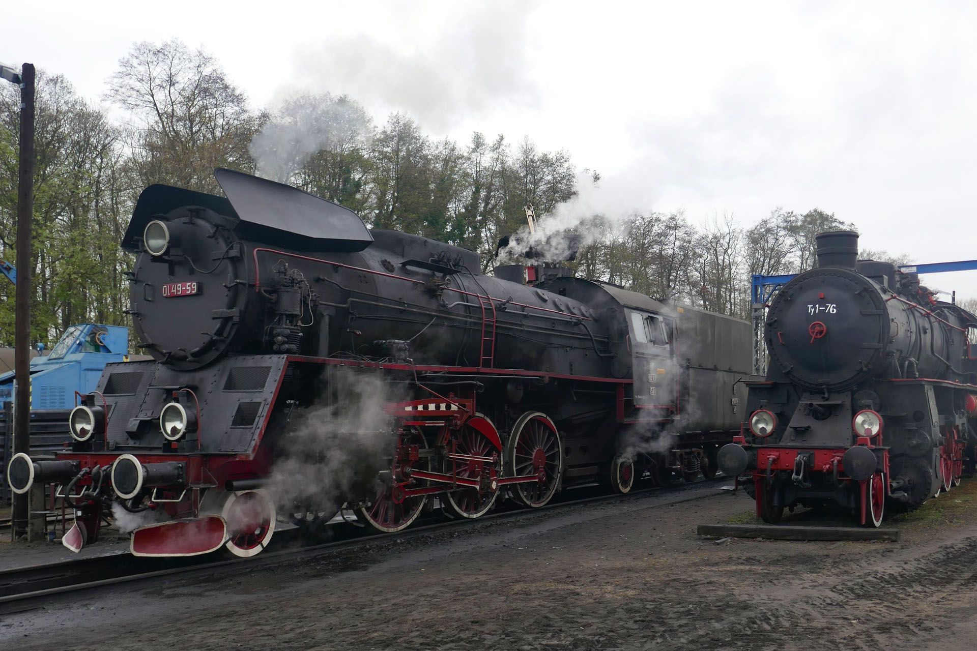Two black steam locomotives with red details, emitting steam, parked on tracks outdoors.