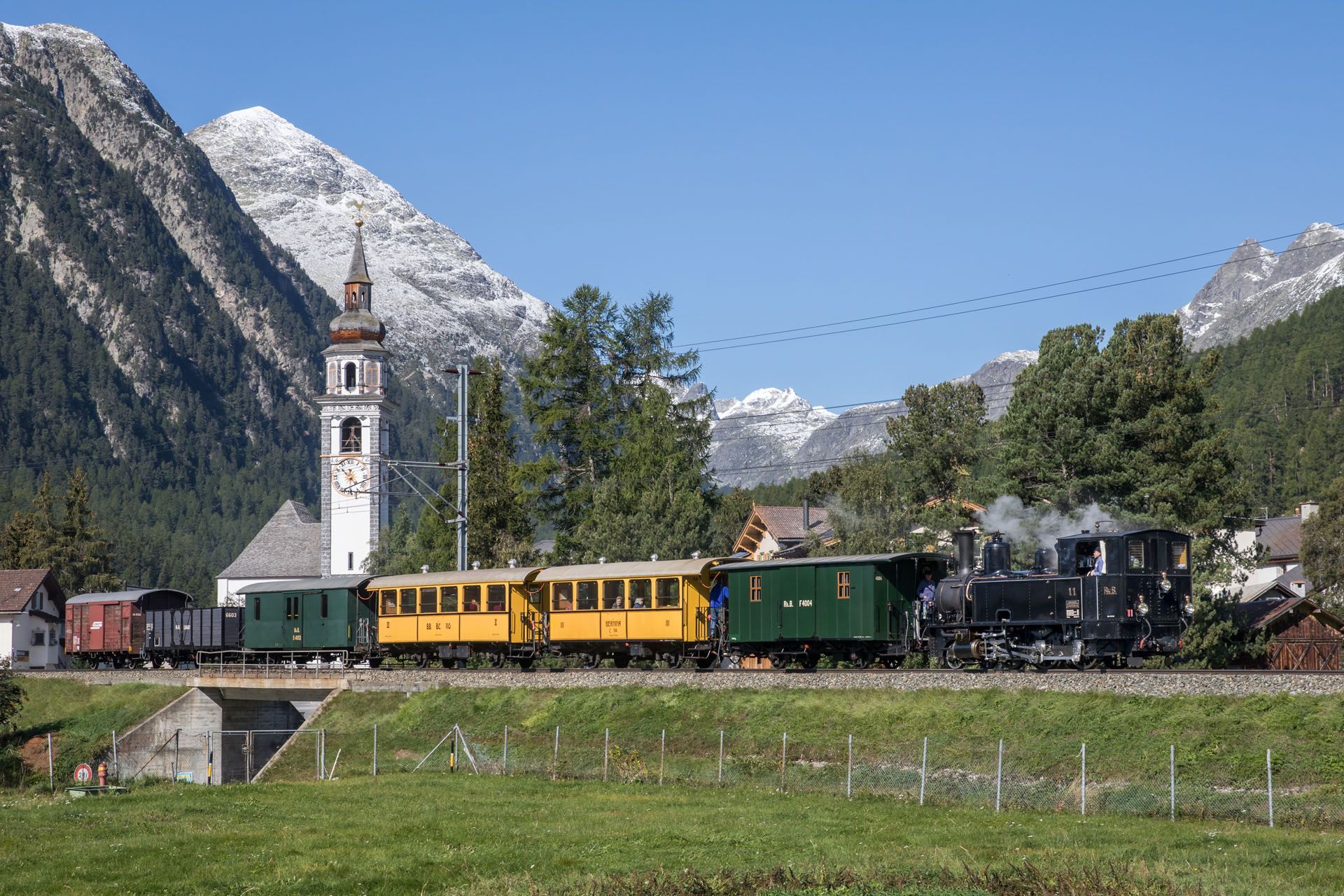 Steam train traveling past a church in a mountain village; yellow and green carriages.