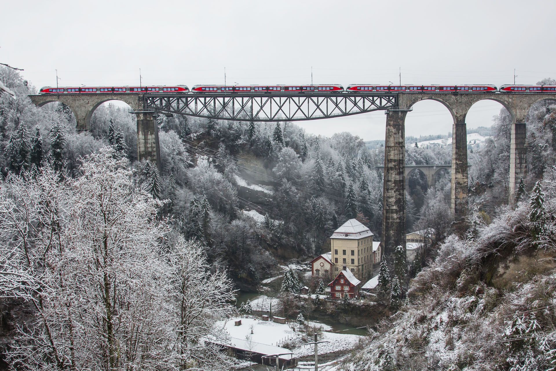 Train crossing a snowy viaduct bridge over a ravine. Winter scene with trees and buildings.