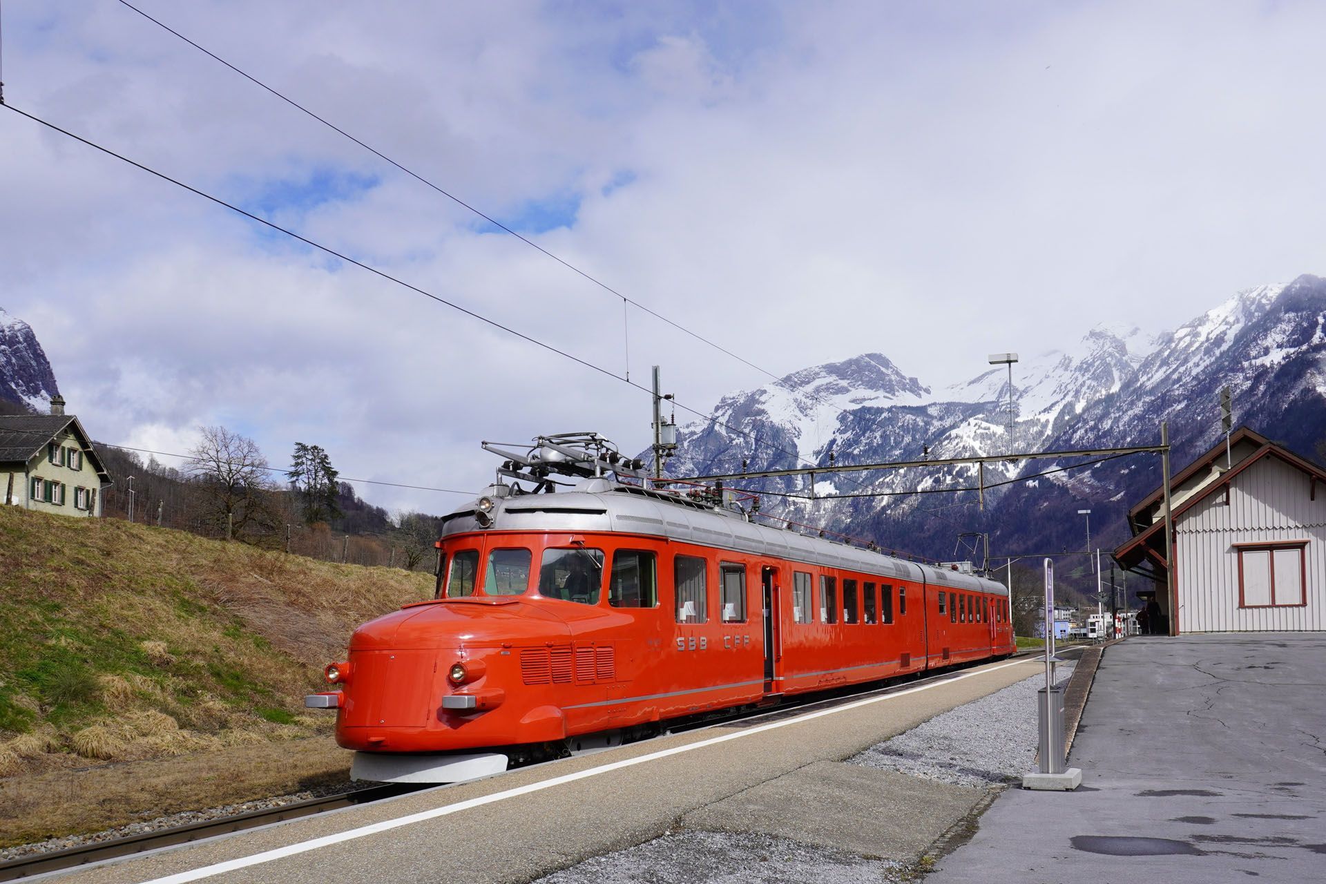Red electric train on tracks near a station, mountains and cloudy sky in background.